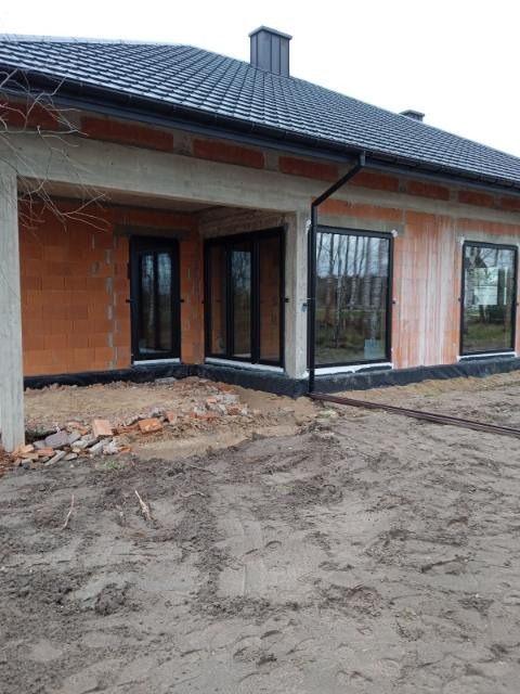 Surowy stan budynku z zamontowanymi czarnymi oknami trzyszybowymi, profil Veka Softline 82, widoczne niedokończone mury i ziemia przed budynkiem.