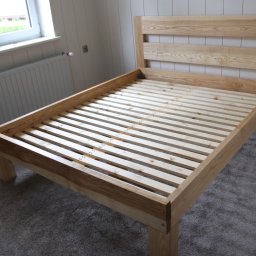 Woodluck - Surowa rama łóżka z jasnego drewna z listwowym stelażem, widoczna w pokoju z szarym dywanem i białymi panelami ściennymi.