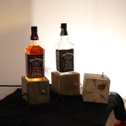 Woodluck - Dwie butelki Jack Daniel's umieszczone na drewnianych podstawkach z wbudowanymi włącznikami, tworzące lampki nocne. Jedna butelka pełna, druga pusta. Trzecia, surowa, drewniana kostka z włącznikiem...