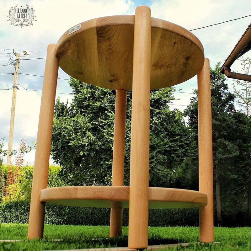 Drewniany, okrągły stolik pomocniczy z dwoma blatami, wykonany z jasnego drewna, ustawiony na trawie, z logo producenta w lewym górnym rogu.