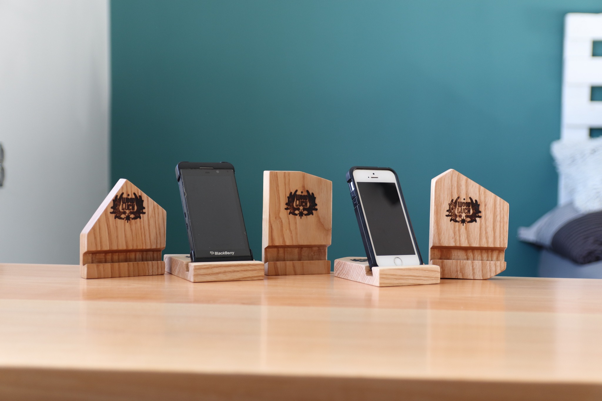 Ręcznie robione, drewniane podstawki pod telefony z wygrawerowanym logo, ustawione na drewnianym blacie na tle dwukolorowej ściany.