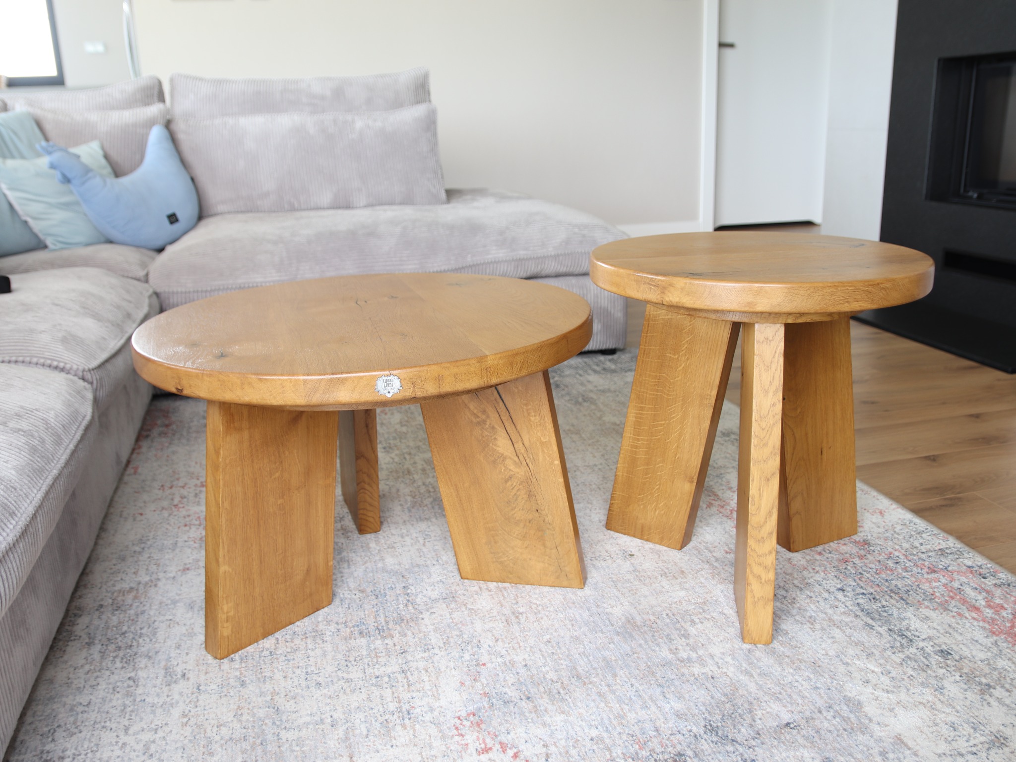 Dwa okrągłe stoliki kawowe z jasnego drewna o minimalistycznym designie, z widocznymi słojami, ustawione na wzorzystym dywanie w salonie z szarą sofą i czarnym kominkiem w tle.