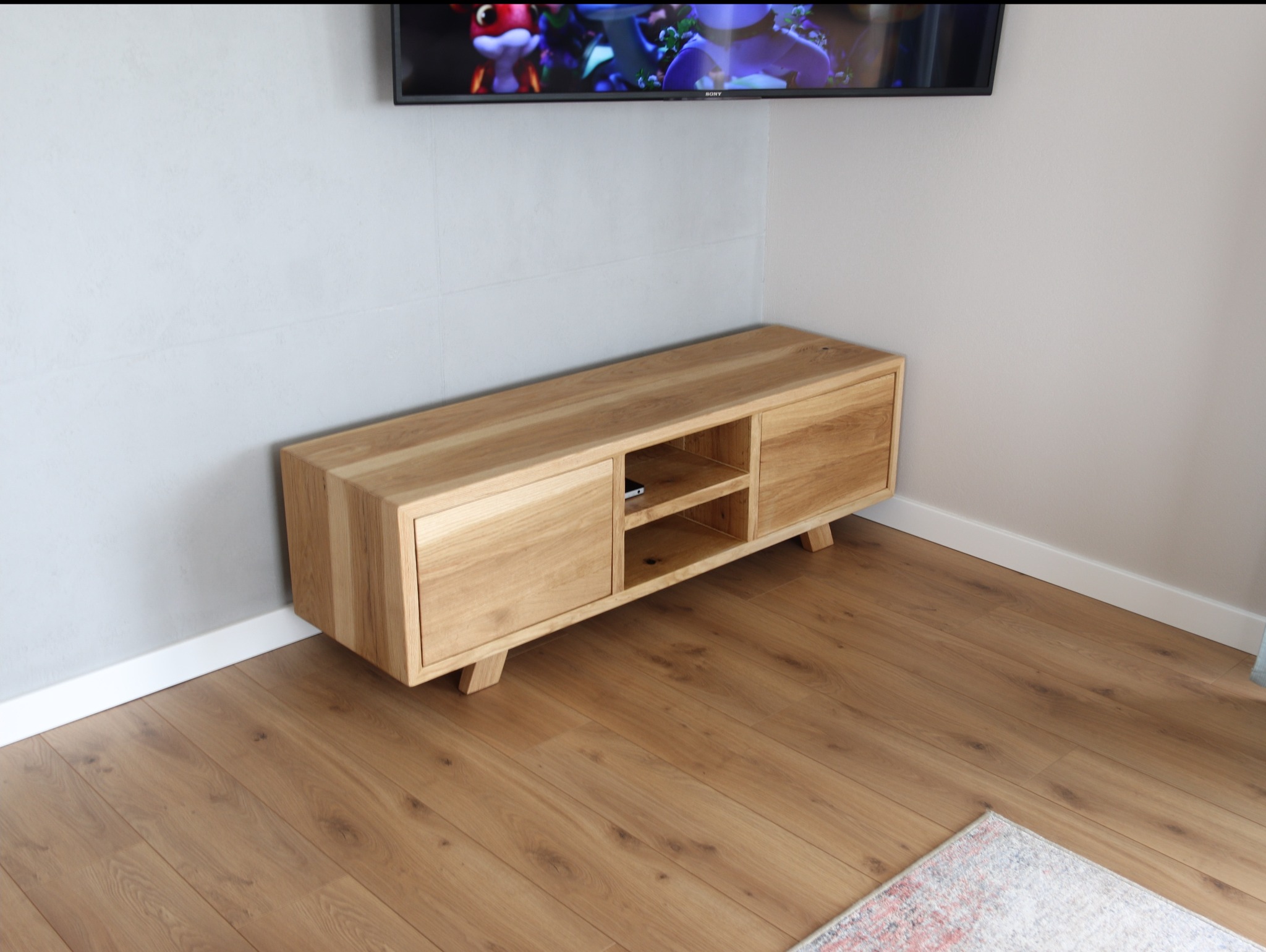 Drewniana szafka RTV z jasnego drewna, z dwiema zamykanymi szafkami i otwartą półką, ustawiona na drewnianej podłodze pod telewizorem w rogu pokoju.