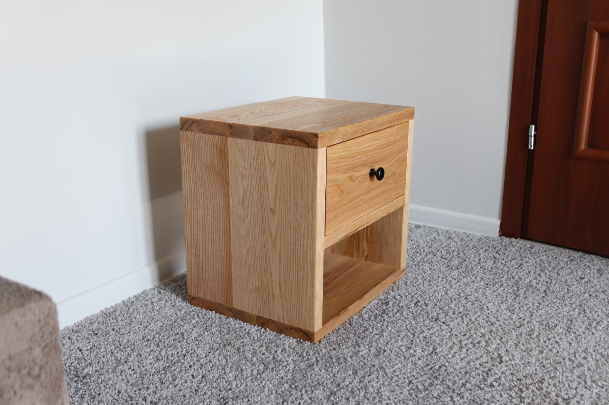 Drewniana szafka nocna z jedną szufladą i otwartą półką na tle białej ściany i brązowych drzwi, stojąca na szarym dywanie.