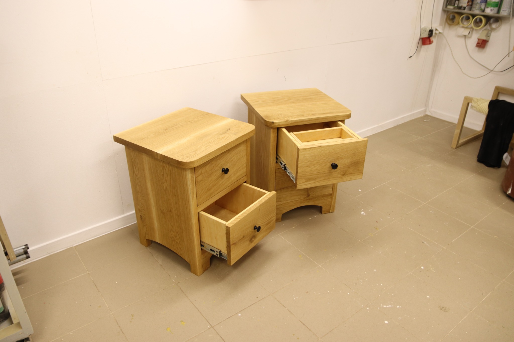 Dwie drewniane szafki nocne z otwartymi szufladami, wykonane z jasnego drewna, widoczne w warsztacie na tle białej ściany i beżowej podłogi z płytek.