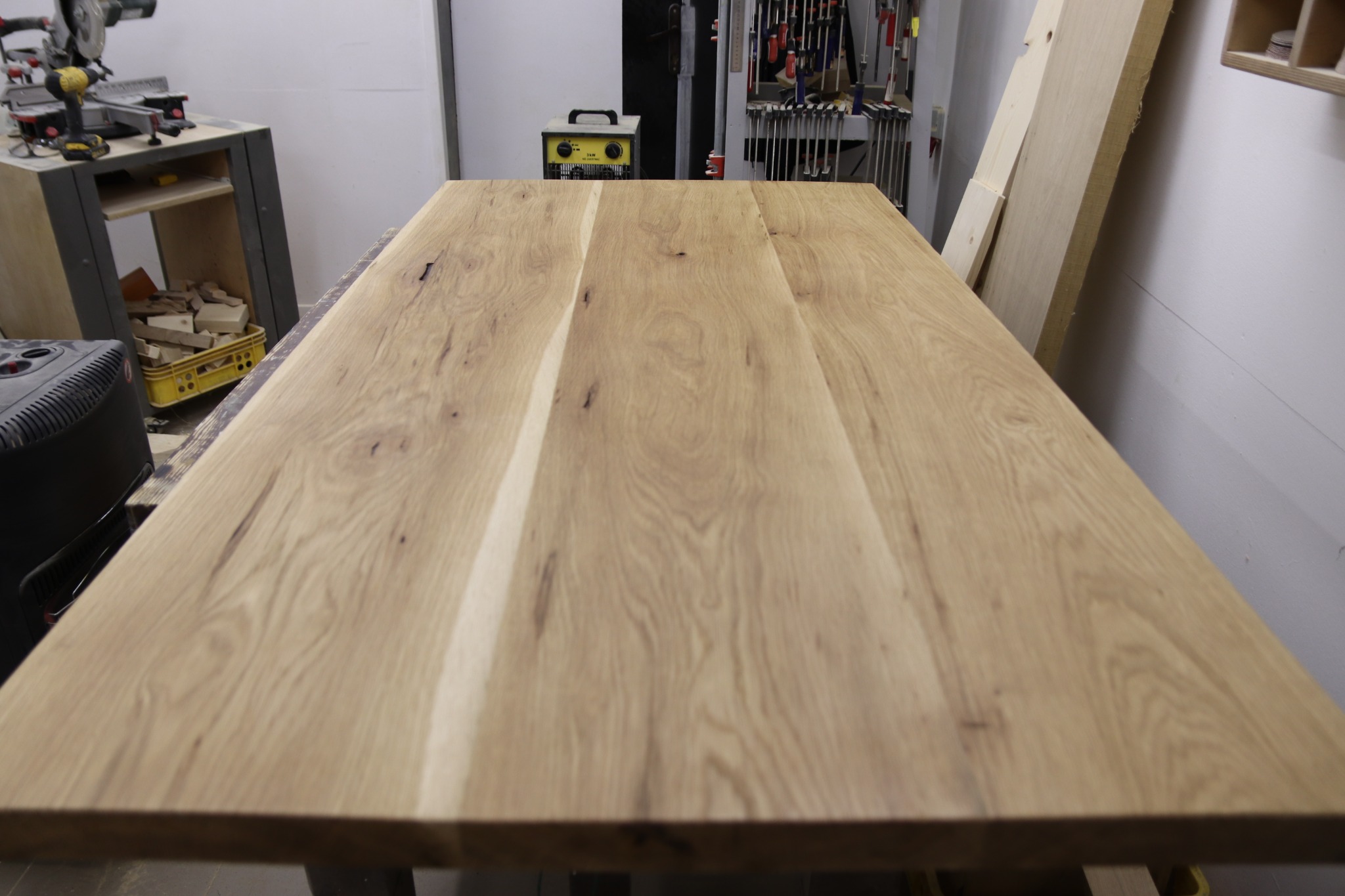 Surowy blat z jasnego drewna z widocznym usłojeniem, ułożony w warsztacie stolarskim z narzędziami i urządzeniami w tle.