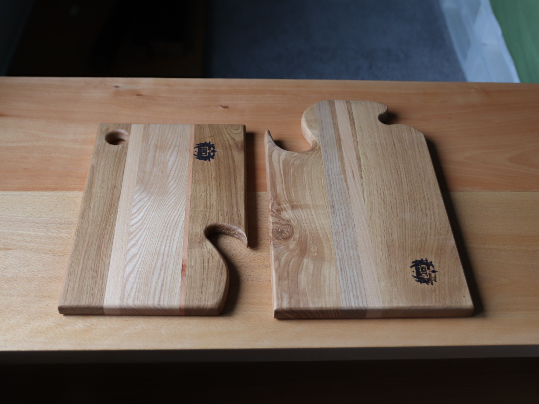 Dwie drewniane deski do krojenia o nieregularnych kształtach, z wypalonym logo, leżące na jasnym drewnianym blacie.