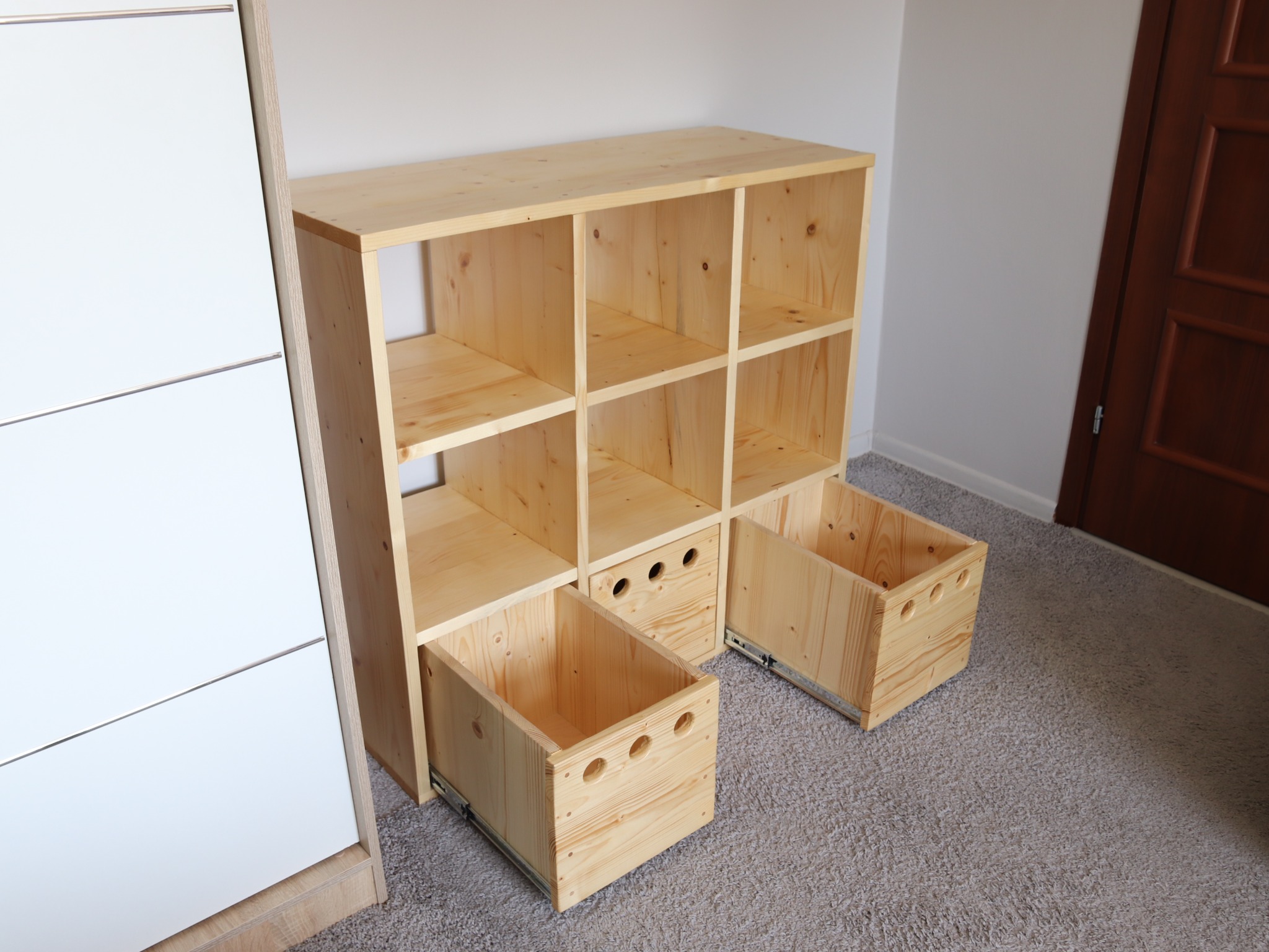 Drewniany regał z otwartymi półkami i dwoma wysuwanymi szufladami na metalowych prowadnicach, wykonany z jasnego drewna sosnowego, na tle szarej wykładziny.