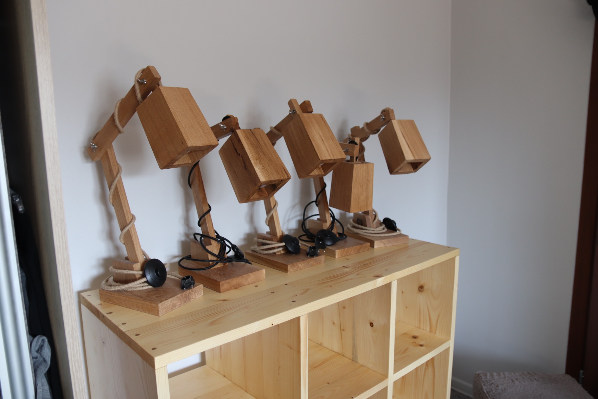 Cztery identyczne lampki biurkowe z jasnego drewna, z elementami sznurka i czarnymi kablami, stojące na drewnianej szafce z otwartymi półkami.