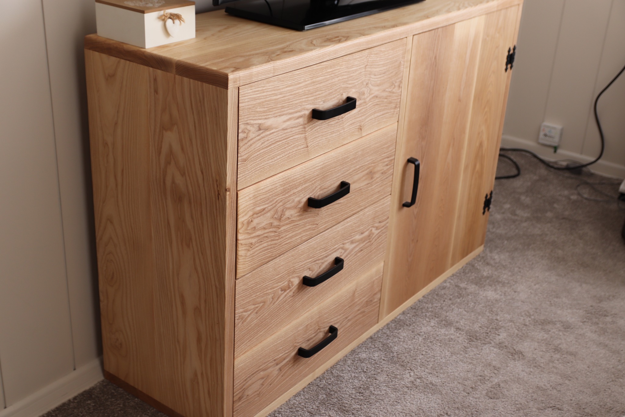 Drewniana komoda z trzema szufladami i szafką z czarnymi uchwytami, stojąca na szarym dywanie, z dekoracyjnym pudełkiem na blacie.