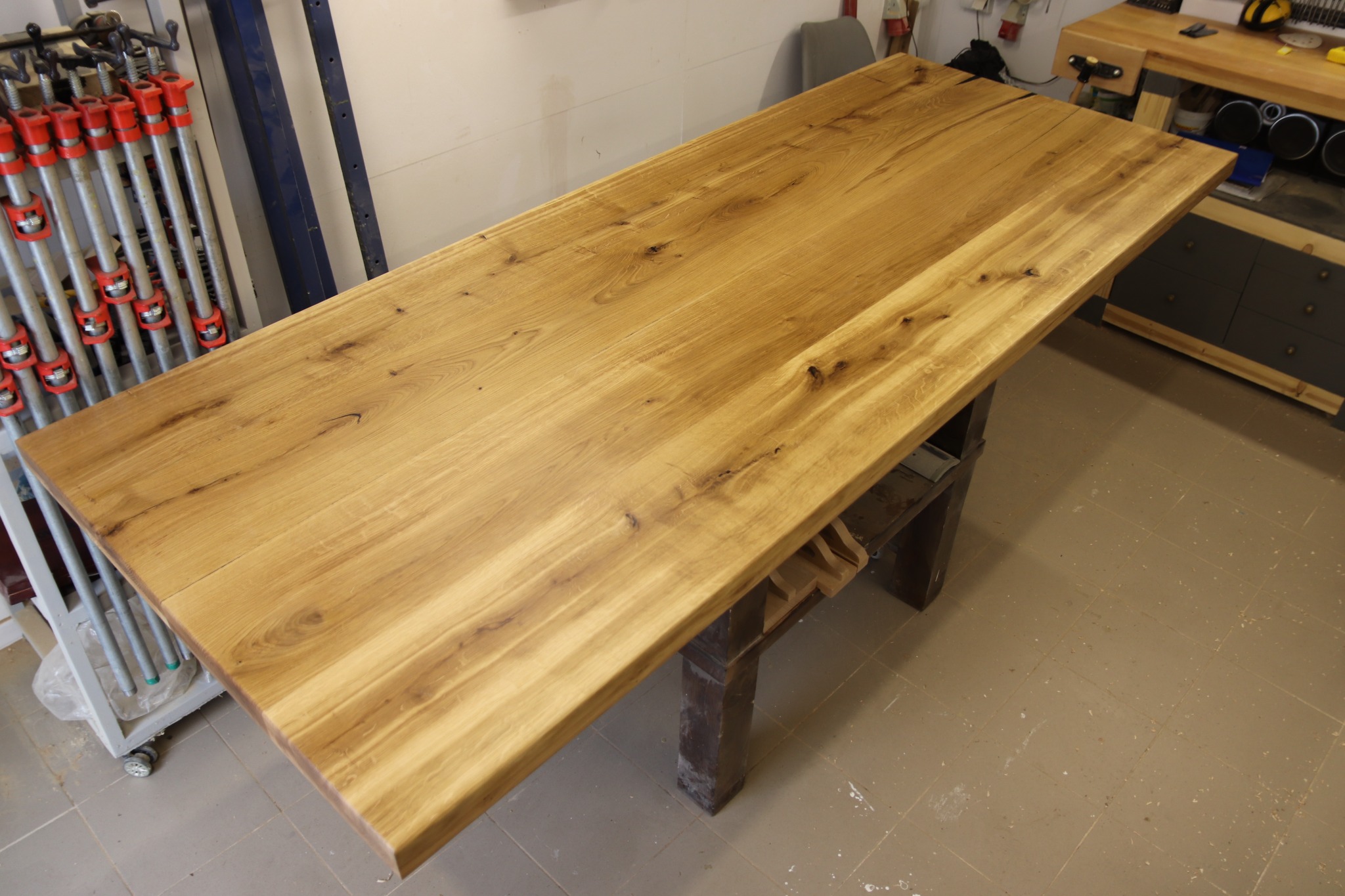 Gotowy blat z jasnego drewna z widocznymi słojami, oparty na ciemnej, drewnianej konstrukcji w warsztacie stolarskim, obok stoją ściski stolarskie.