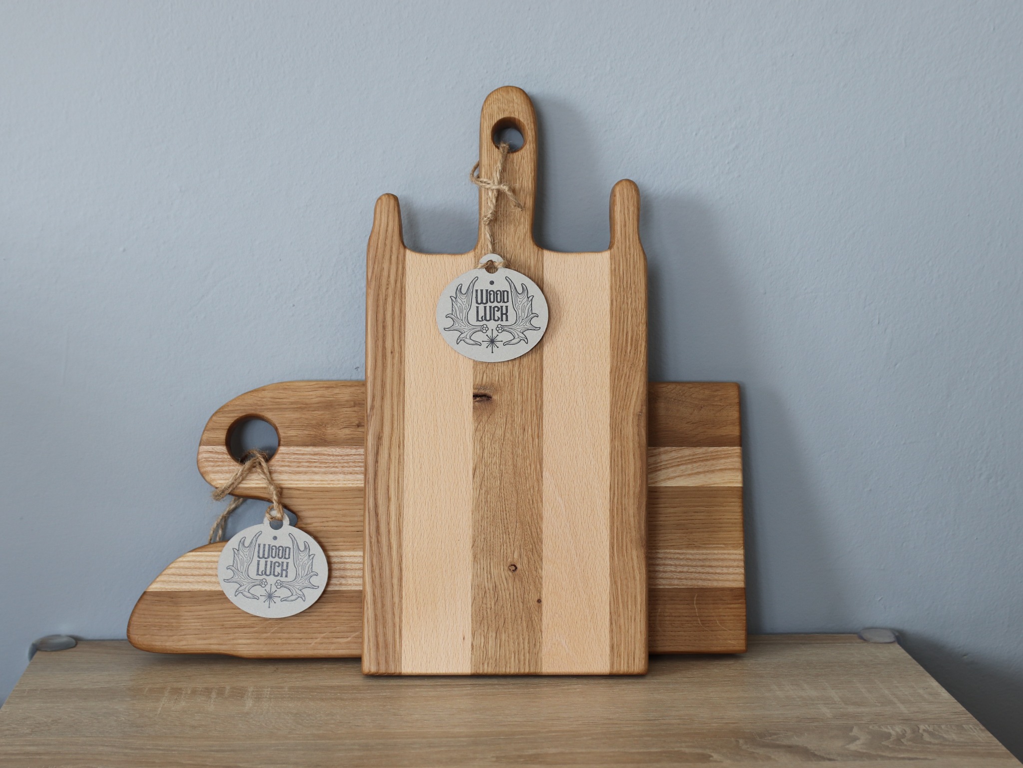 Dwie drewniane deski do krojenia z zawieszkami marki 'Wood Luck' na blacie z drewnopodobnego materiału, na tle szarej ściany.