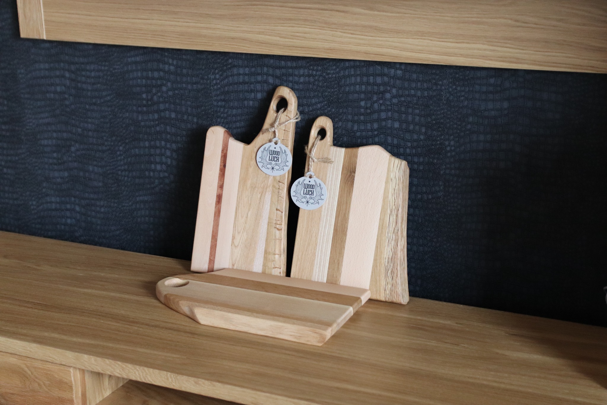 Trzy drewniane deski do krojenia z zawieszkami 'Wood Luch' na blacie z jasnego drewna, na tle ciemnej ściany o fakturze skóry.
