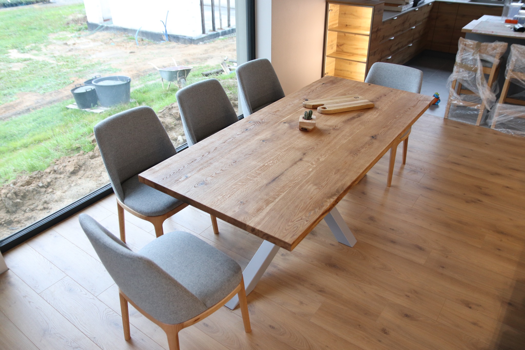 Drewniany stół z szarymi krzesłami w jasnym wnętrzu, widok na plac budowy przez okno, na blacie deski do krojenia i mała roślina w doniczce.