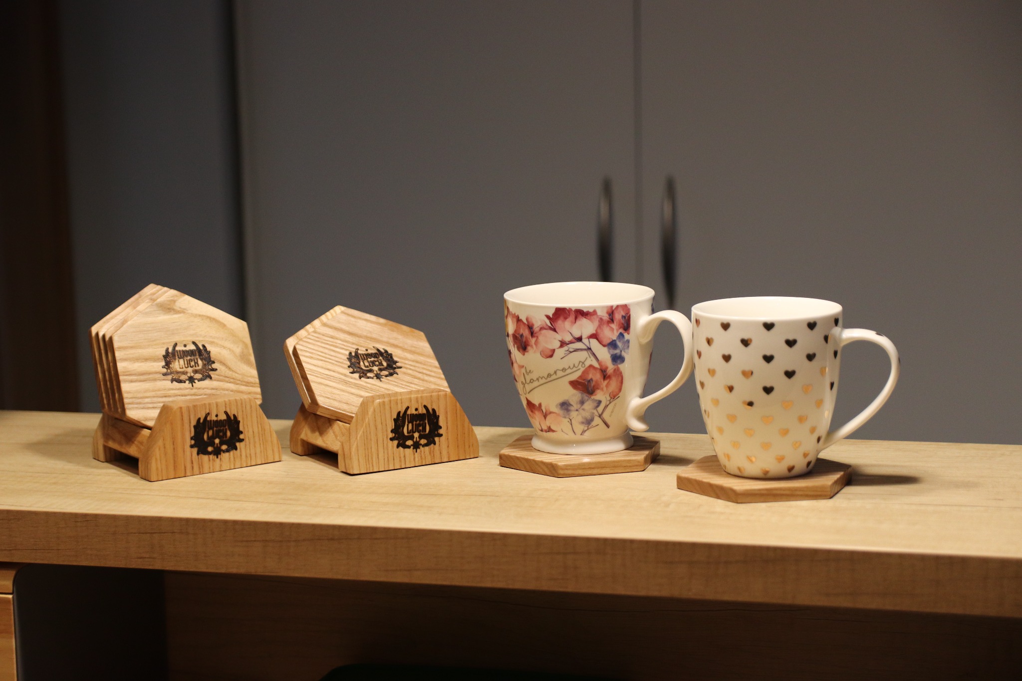 Dwa komplety drewnianych podkładek pod kubki z wypalonym logo firmy 'Wood Luck', ustawione na blacie; kubki z motywami kwiatowymi i serduszkami.