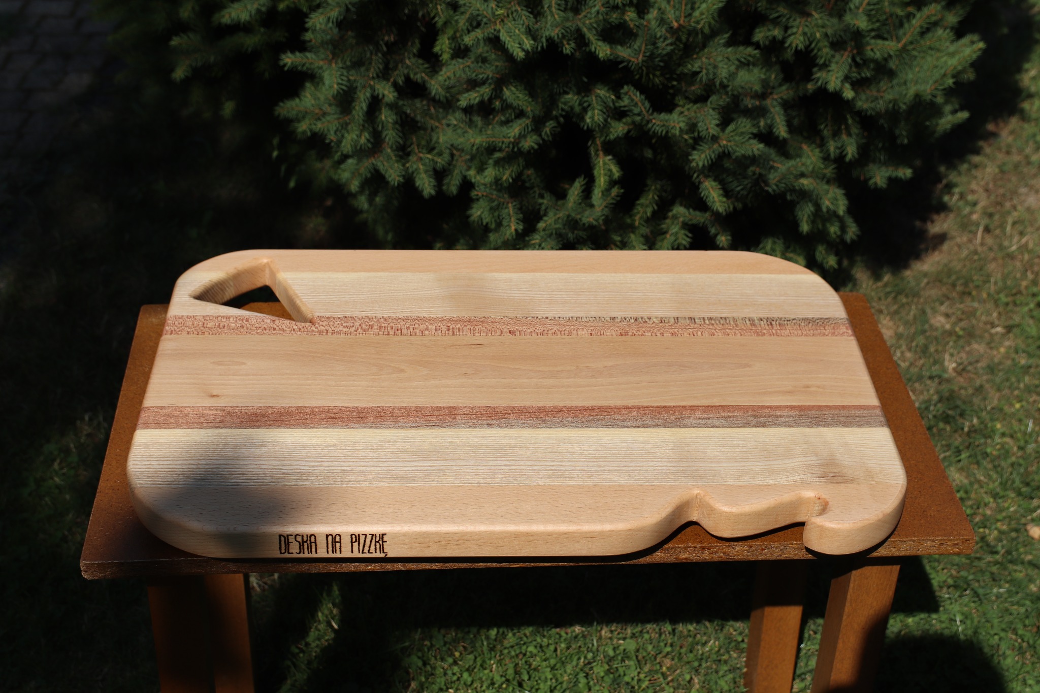 Drewniana deska do pizzy z uchwytem i wygrawerowanym napisem 'DESKA NA PIZZKE', ułożona na brązowym stoliku na tle zielonego trawnika i krzewu iglastego.