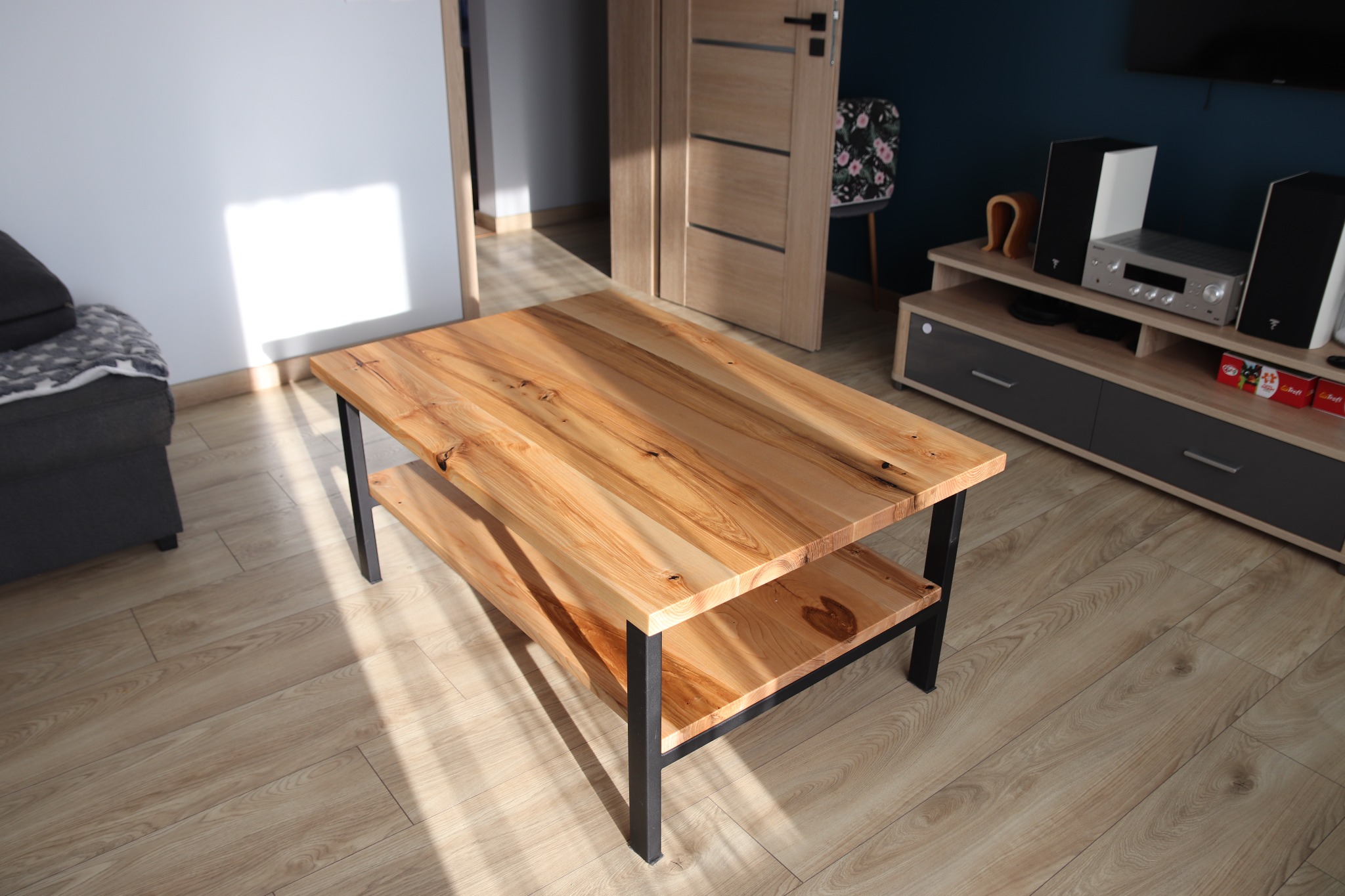 Stolik kawowy z blatem z jasnego drewna o wyraźnym usłojeniu, na czarnych metalowych nogach, z dodatkową półką, w nowoczesnym salonie z szarą sofą i szafką RTV.