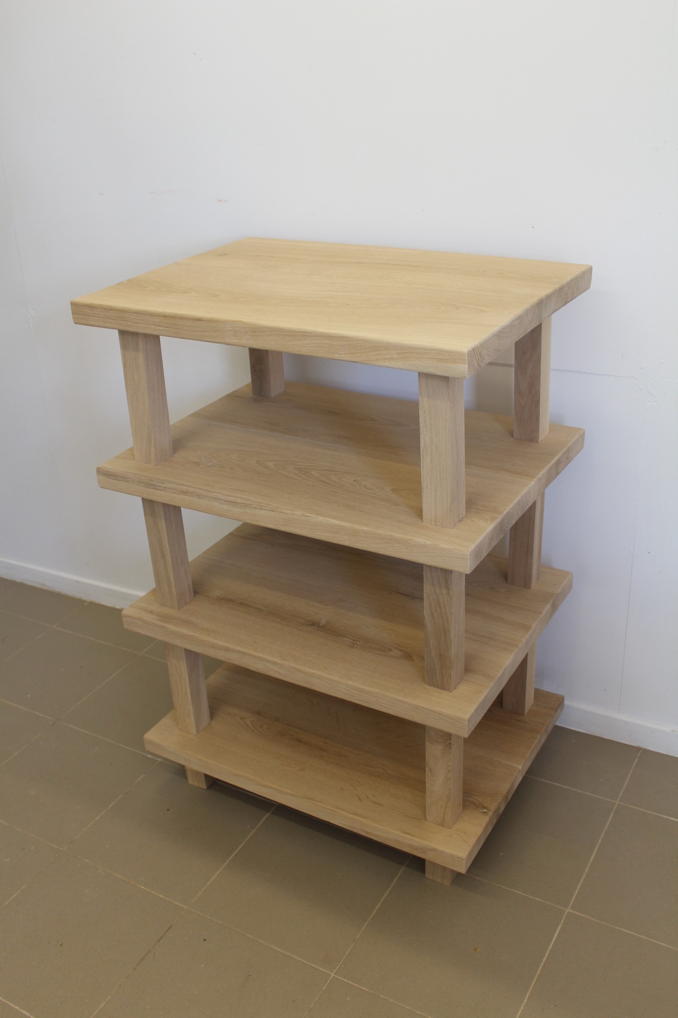 Trójpoziomowy regał z jasnego drewna o minimalistycznym designie, z grubymi, kwadratowymi nogami, na tle białej ściany i szarej podłogi.