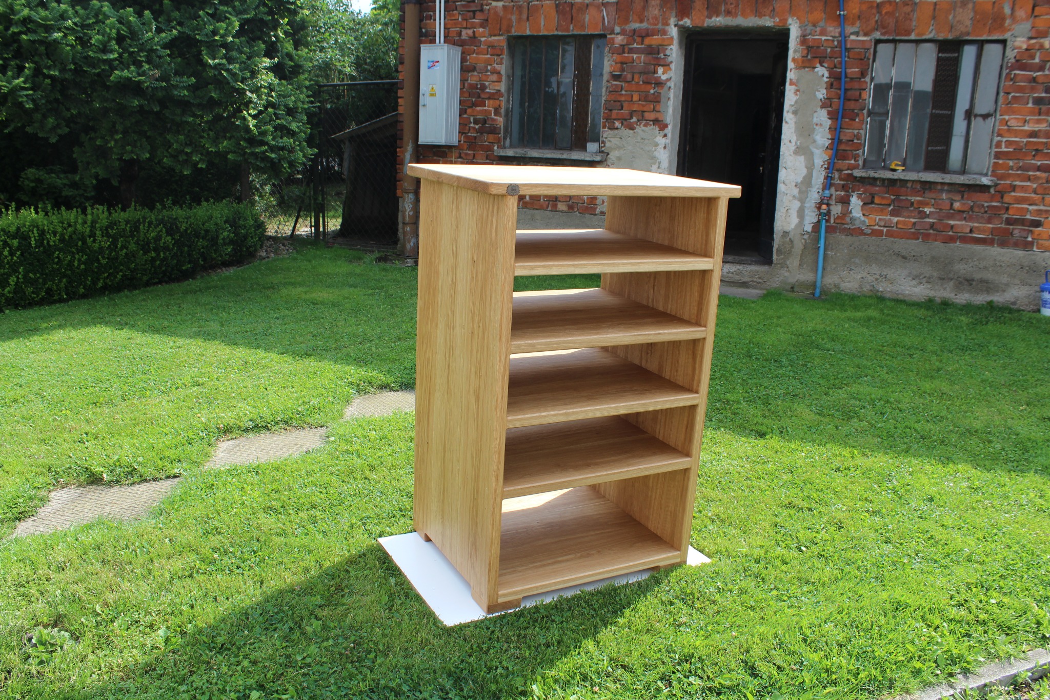 Drewniana szafka z półkami ustawiona na trawie przed ceglanym budynkiem z oknami.