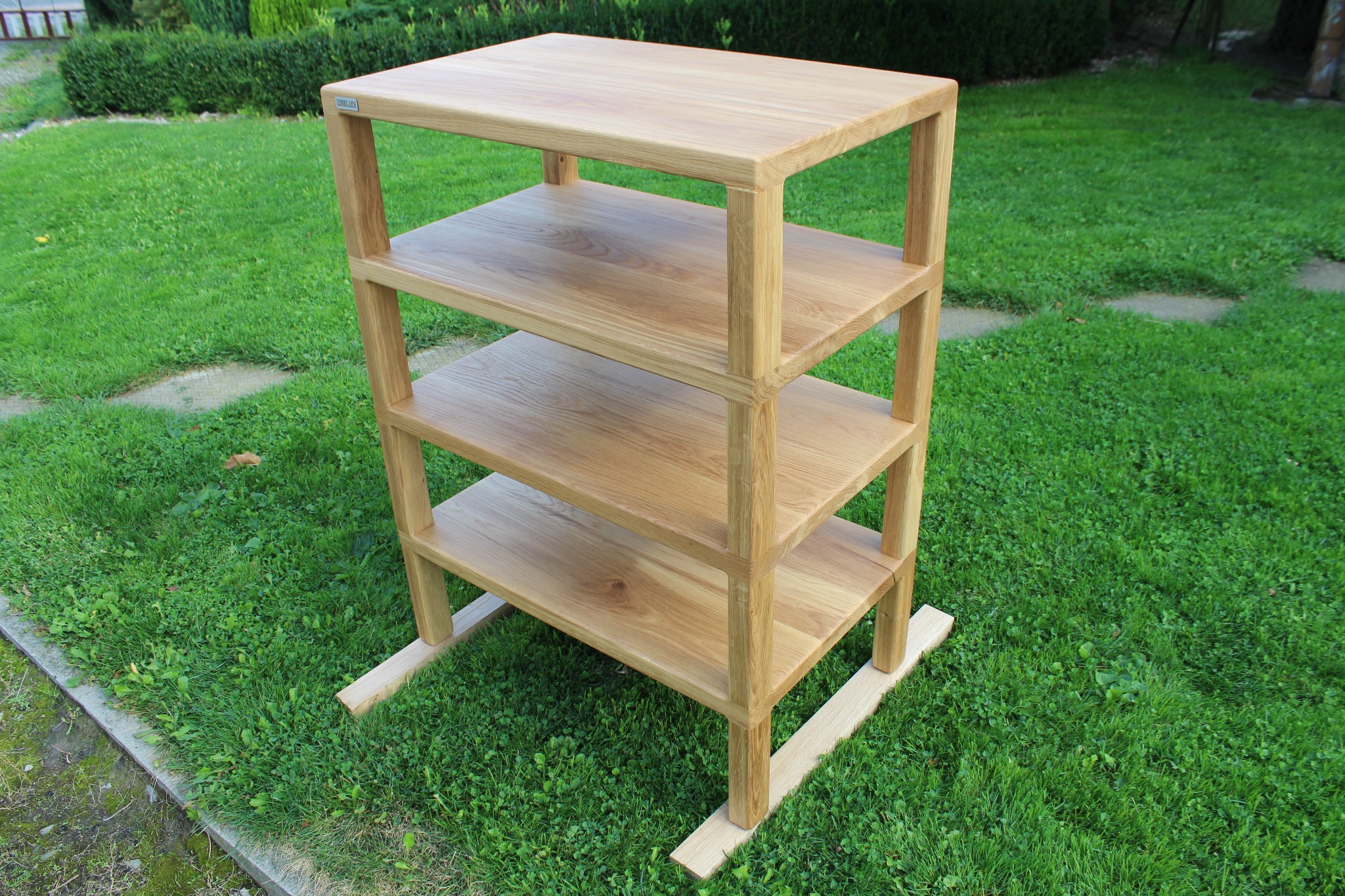 Drewniany regał z czterema półkami, wykonany z jasnego drewna, ustawiony na trawie w ogrodzie. Widoczne detale łączeń i naturalna faktura drewna.