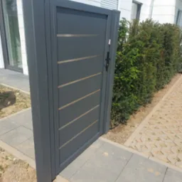 Nowoczesna, grafitowa furtka ogrodowa z poziomymi, jasnymi akcentami, zamontowana przy ścieżce z betonowych płyt i żywopłocie.