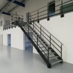 Wewnętrzne metalowe schody z balustradami ze stali nierdzewnej w hali produkcyjnej lub magazynie, widok z dołu na konstrukcję z perforowanymi stopniami i czarną ramą.