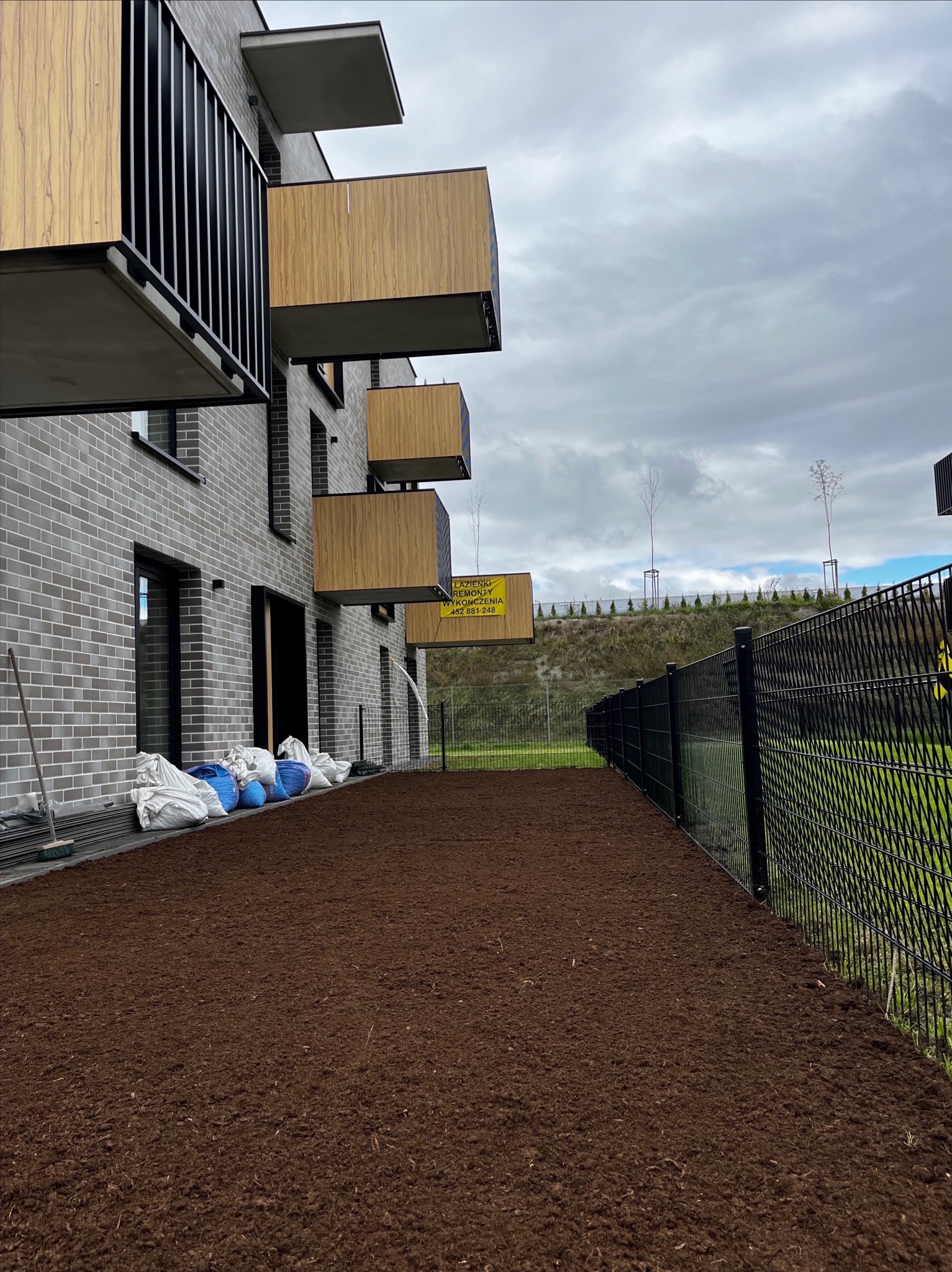 Przygotowany grunt pod trawnik przy nowoczesnym budynku z balkonami. Worki z ziemią i narzędzia ogrodnicze przy ścianie. W tle ogrodzenie i nasadzenia na wzgórzu.