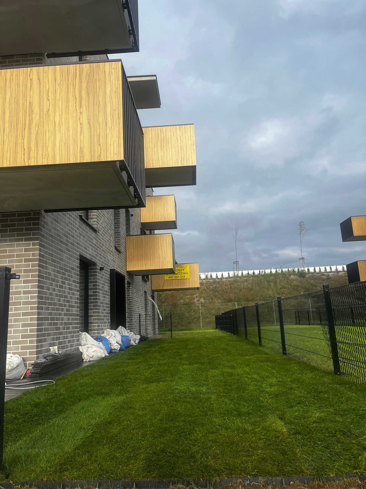 Świeżo założony trawnik z rolki przy nowoczesnym budynku z balkonami w Krakowie. Widoczne worki z ziemią i ogrodzenie.