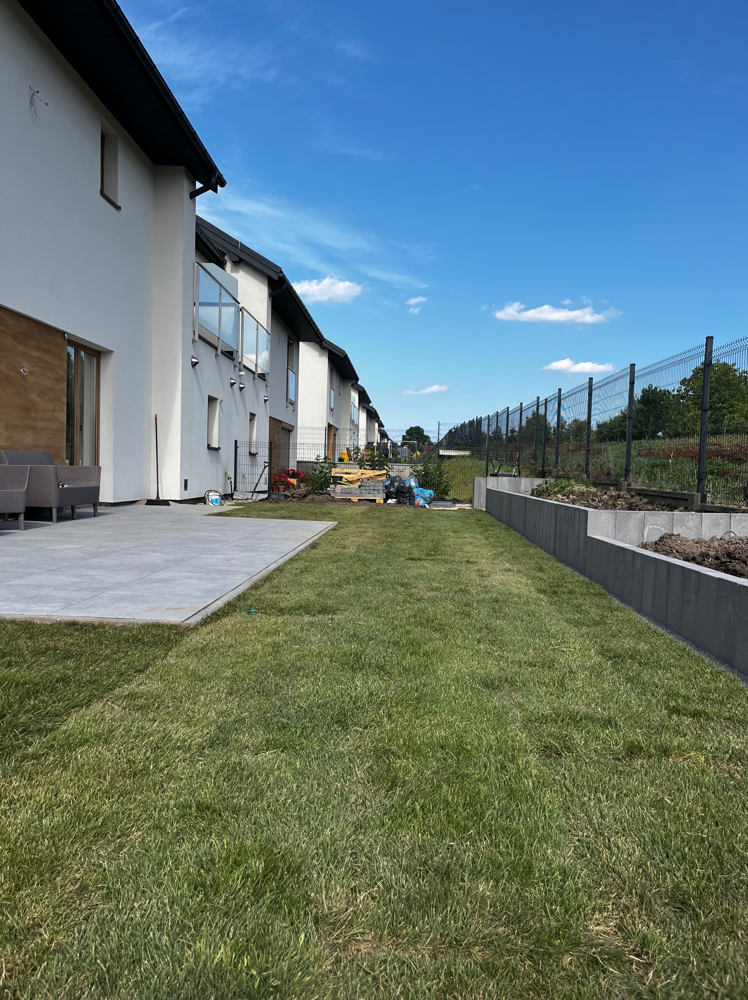 Nowoczesny ogród z trawnikiem, betonowym murem oporowym i siatką ogrodzeniową przy szeregówce, w słoneczny dzień. Widoczne narzędzia ogrodnicze.