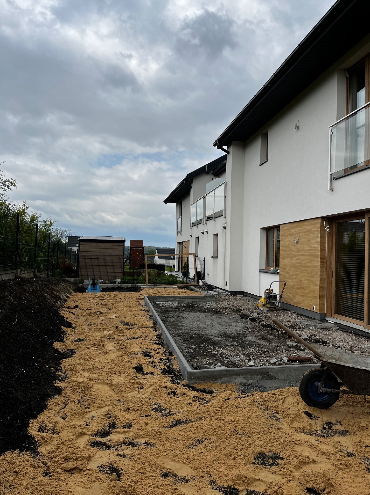 Przygotowanie terenu pod trawnik: wyrównany piasek, obrzeża betonowe, zagęszczarka w tle, nowoczesny dom z balkonami. Etap prac w pochmurny dzień.