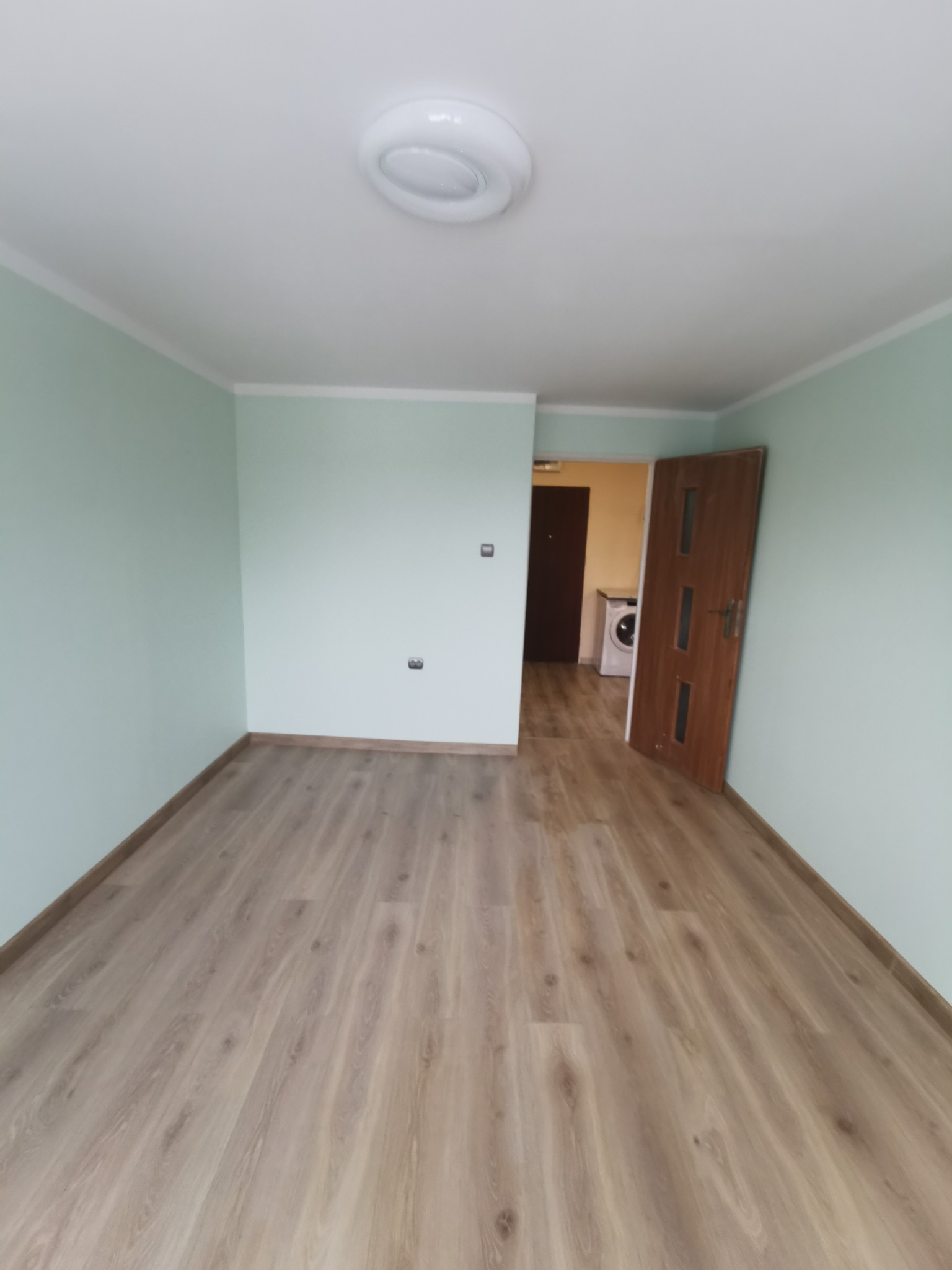 Pusty pokój po remoncie z panelami podłogowymi w odcieniach beżu, świeżo pomalowane ściany na miętowo, widoczna biała lampa sufitowa i otwarte drzwi do pomieszczenia z pralką.