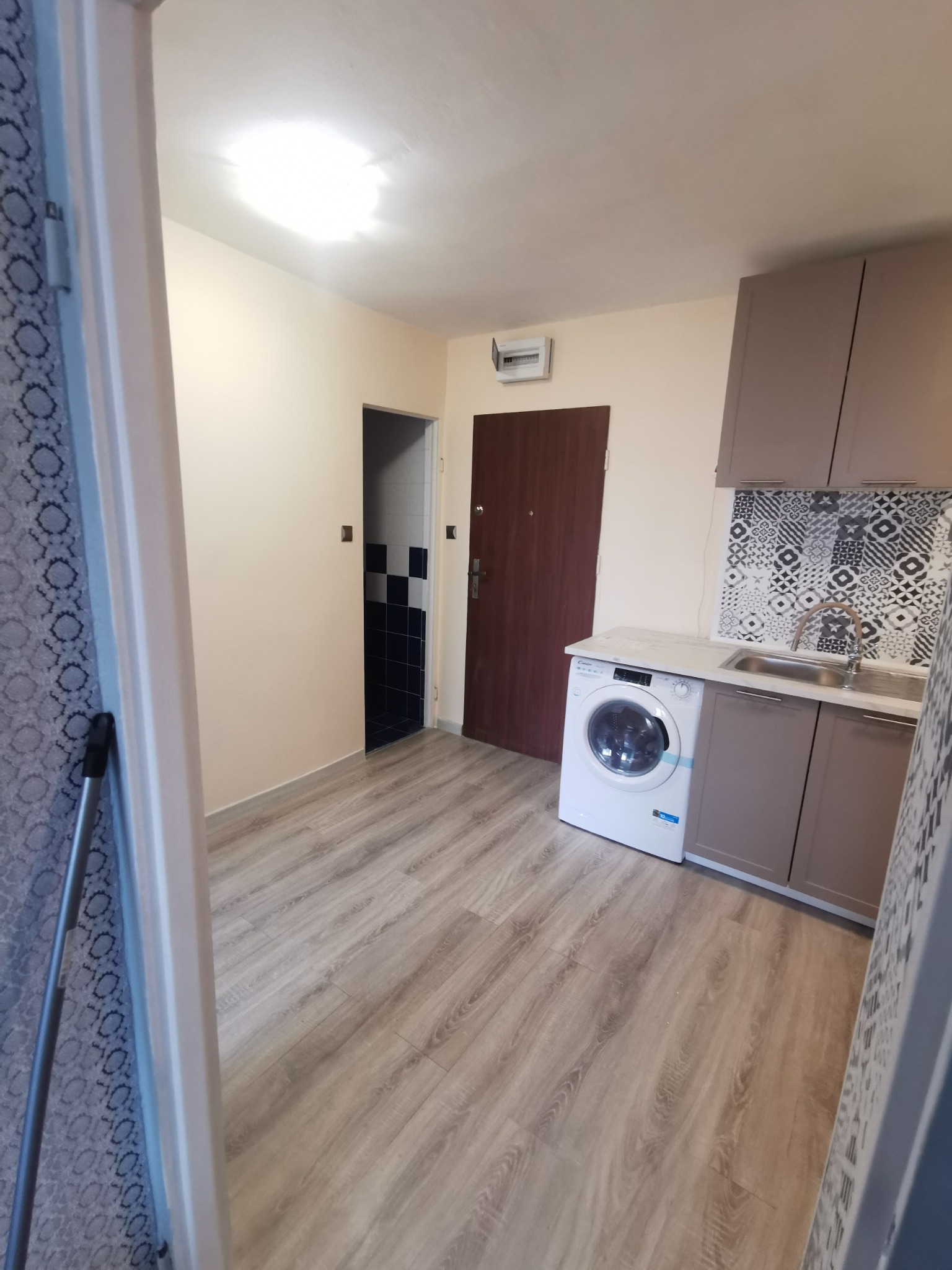 Wykończona kuchnia z pralką pod blatem, szaro-beżowe szafki kuchenne, wzorzysty panel nad blatem, drewnopodobna podłoga, widok na łazienkę z czarno-białymi płytkami.