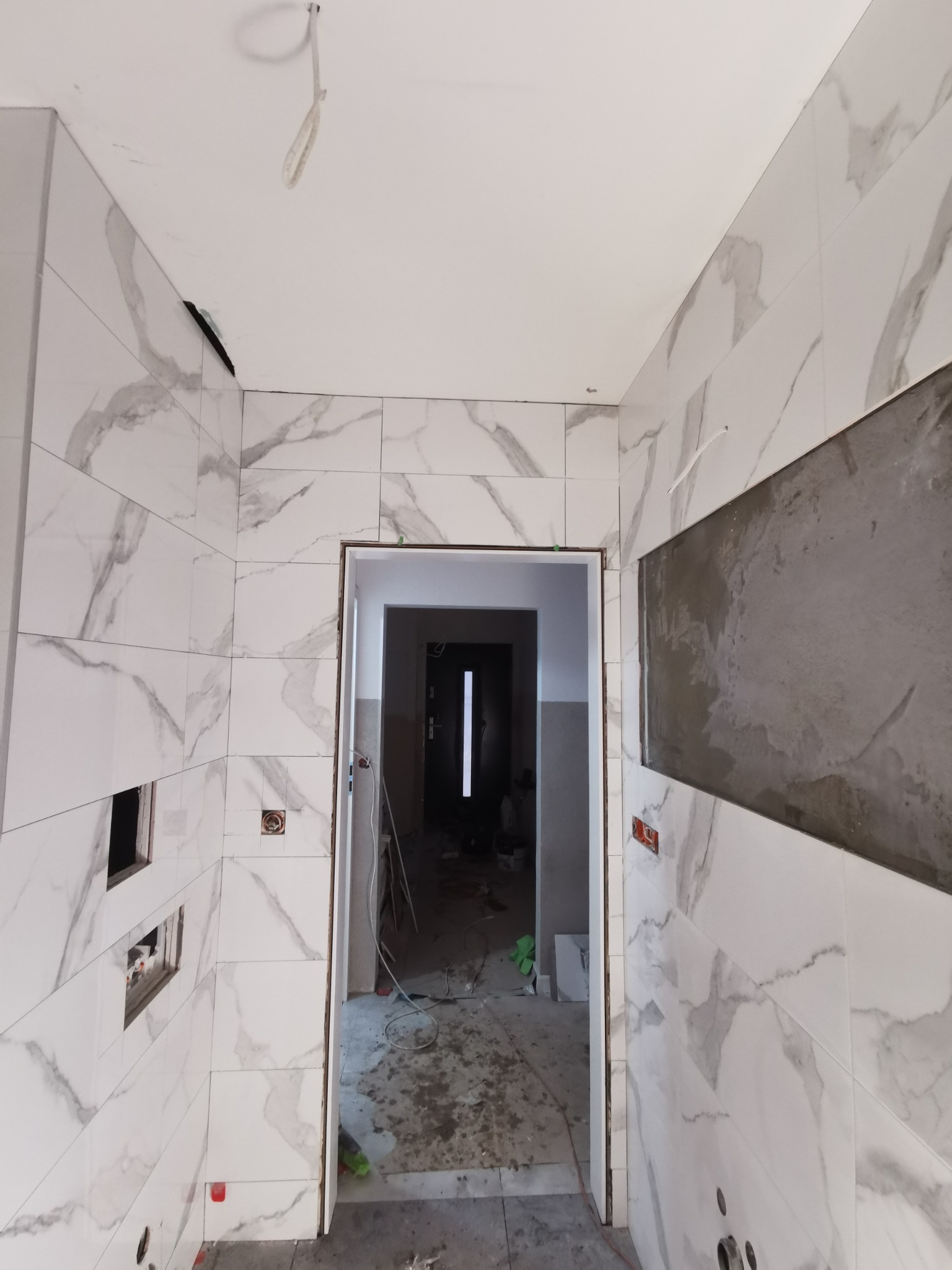 Wnętrze łazienki w trakcie wykańczania, widoczne białe płytki imitujące marmur na ścianach, otwory na instalacje, przygotowane miejsce pod zabudowę oraz widok na korytarz.