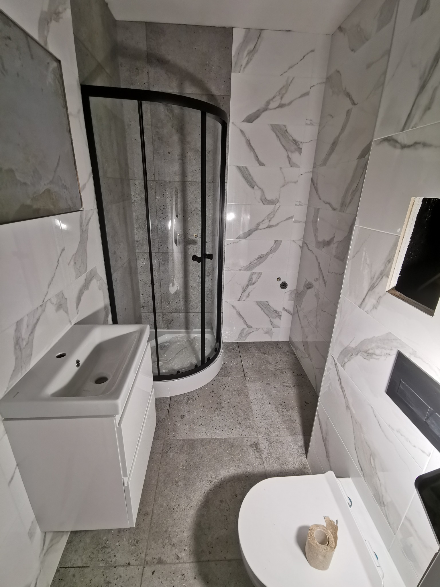 Wykończona łazienka w szaro-białej kolorystyce z kabiną prysznicową z czarnymi profilami, białą umywalką z szafką i toaletą z rolką papieru toaletowego na desce.