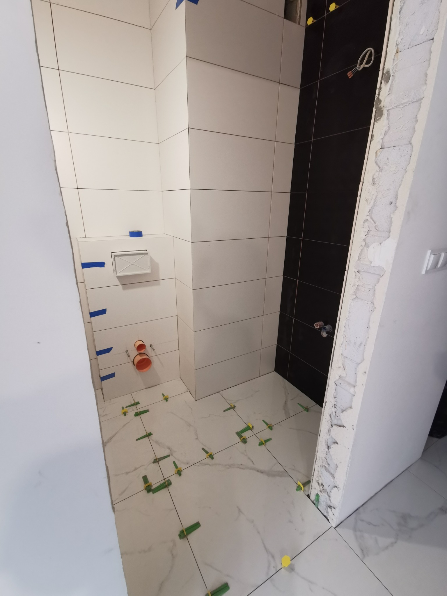 Łazienka w trakcie wykańczania: białe i czarne płytki ścienne, marmurowe płytki podłogowe z zielonymi krzyżykami dystansowymi, widoczne instalacje hydrauliczne i elektryczne, ślady po skuciu tynku.