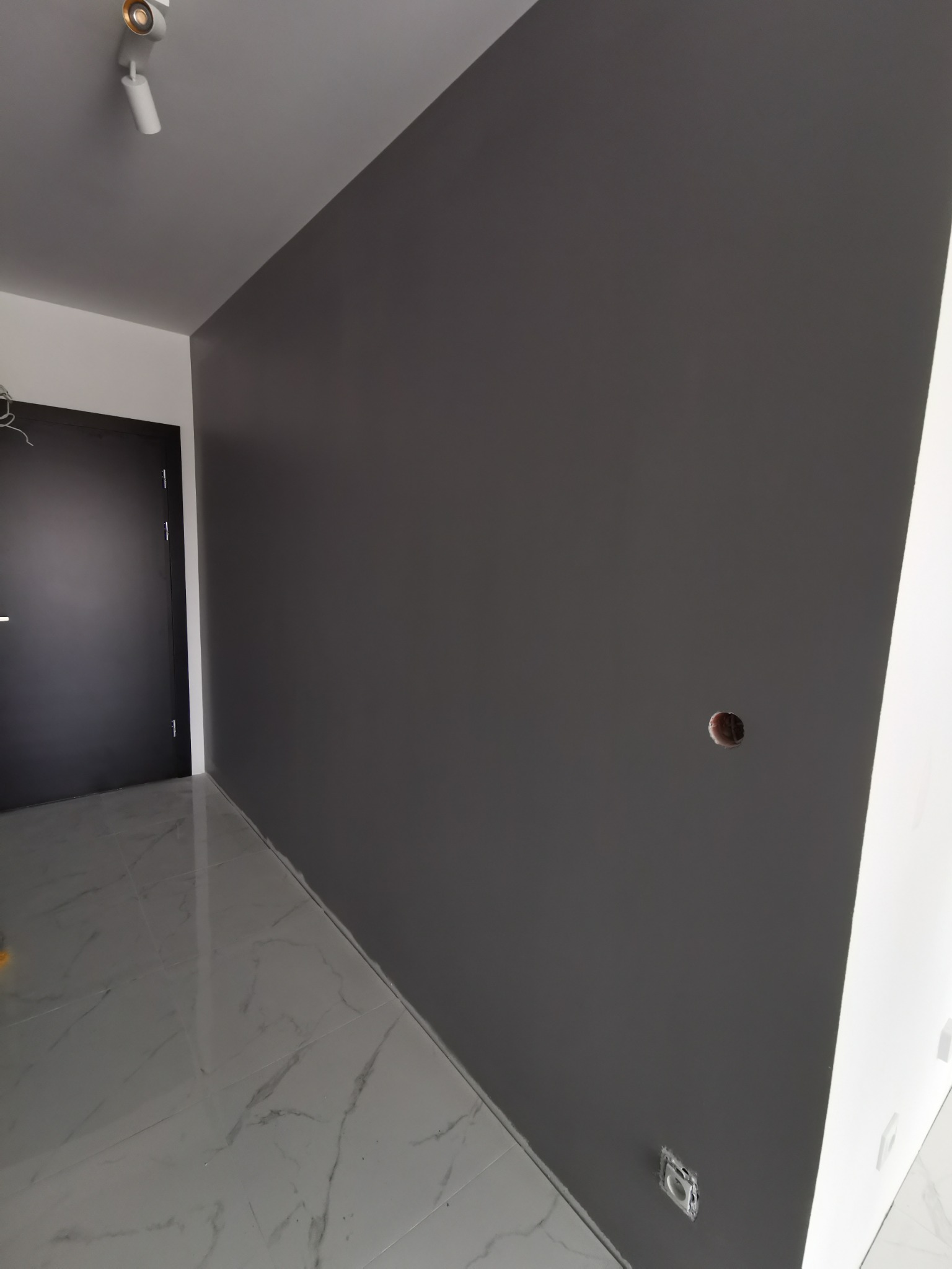 Świeżo pomalowana, ciemnoszara ściana w pomieszczeniu z błyszczącą, marmurkową podłogą, widoczne puszki elektryczne przed montażem gniazdek.