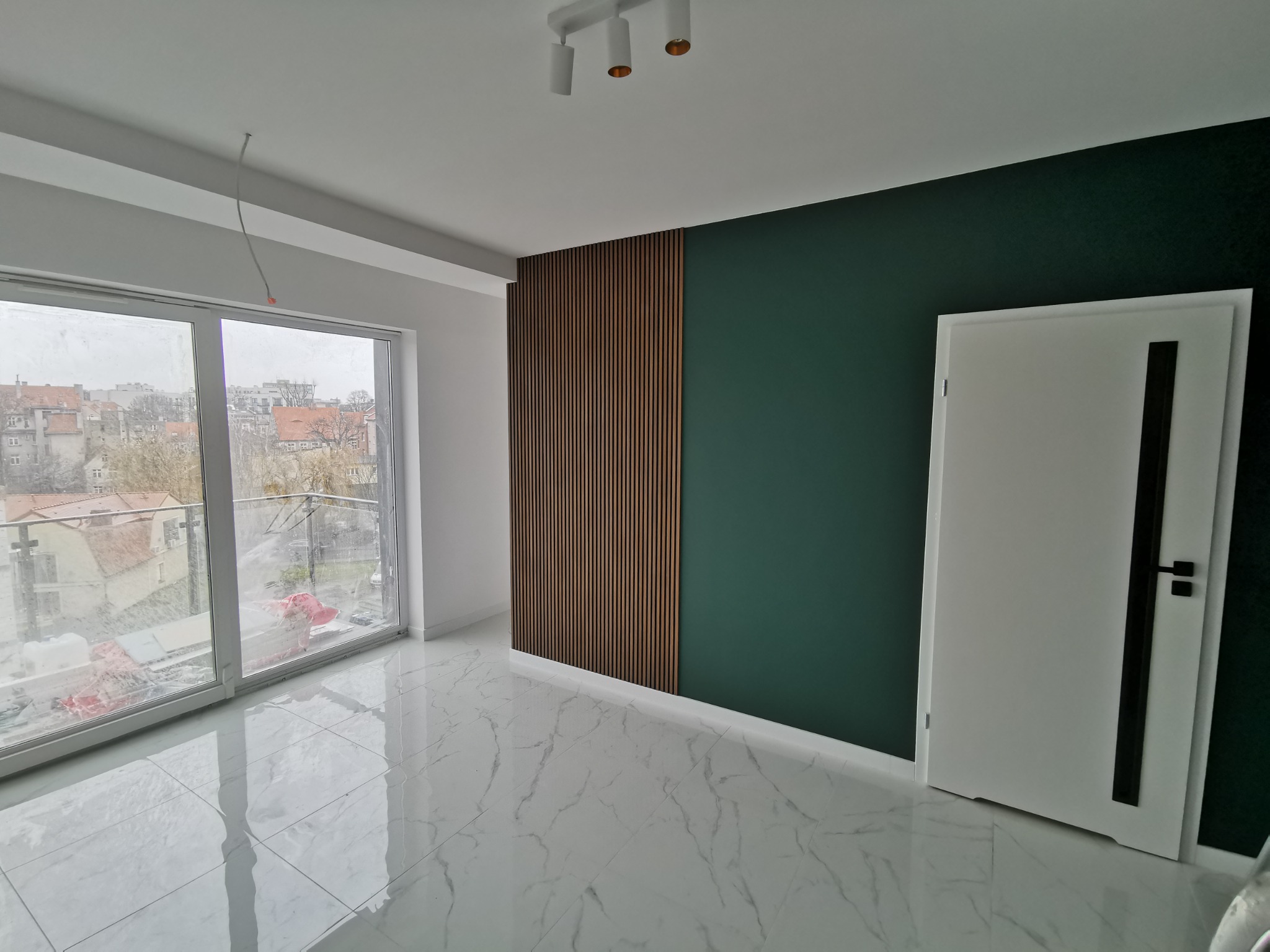 Nowoczesne wnętrze po remoncie: ściana pomalowana na ciemnozielono, panele drewniane pionowe, białe marmurowe płytki podłogowe, białe drzwi z czarnym paskiem, widok na miasto z dużego okna...