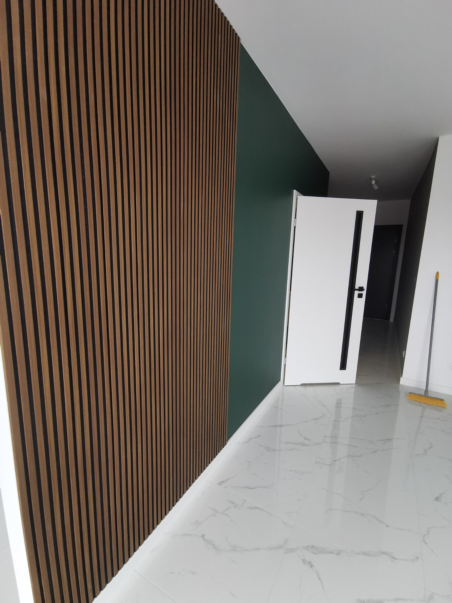 Nowoczesne wnętrze po remoncie: drewniane lamele na ścianie, biała podłoga z marmurowym wzorem, drzwi wewnętrzne z czarnym akcentem i świeżo pomalowana ściana w kolorze butelkowej zieleni.