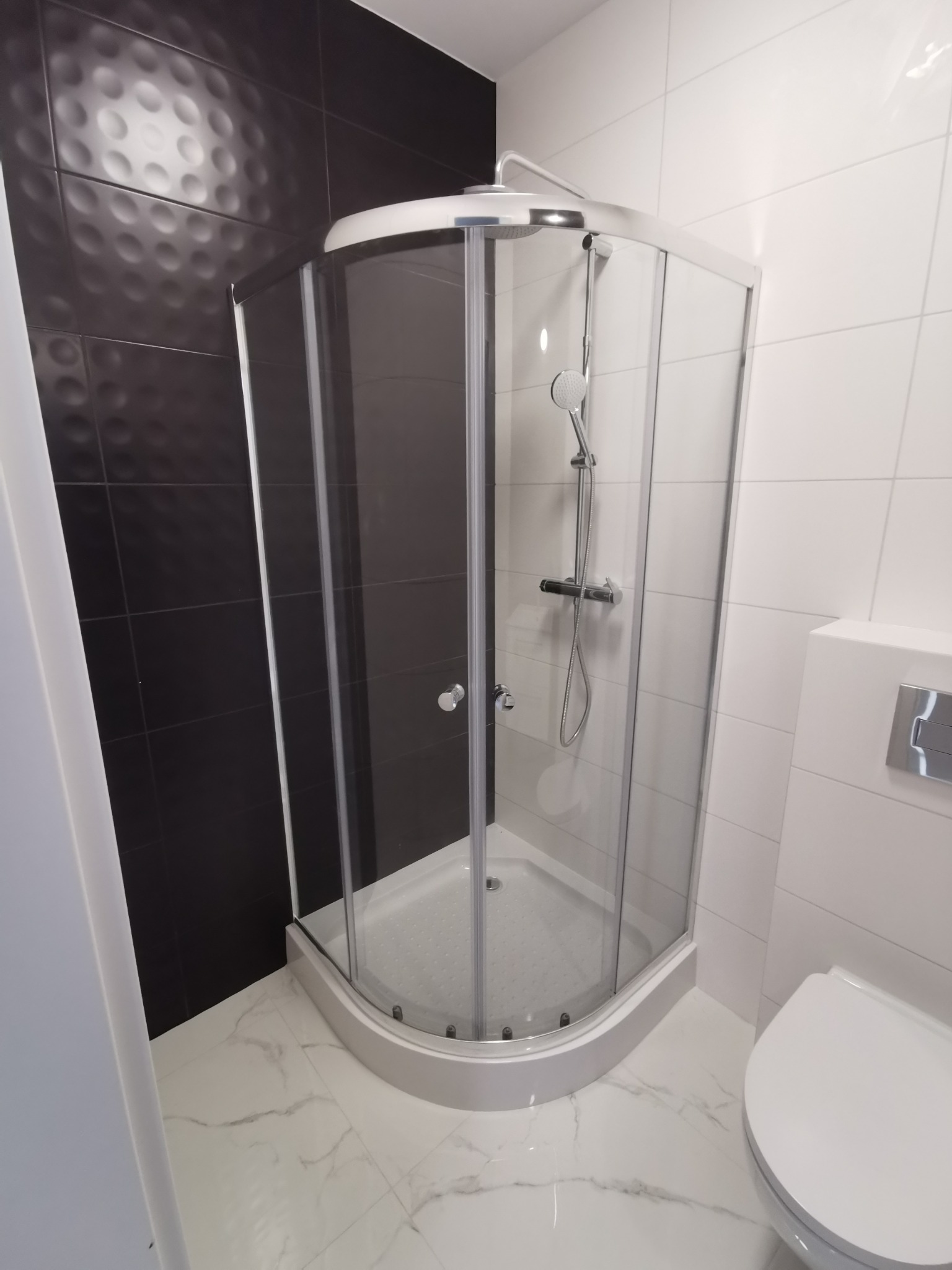 Nowoczesna kabina prysznicowa narożna z chromowaną armaturą na tle czarno-białej łazienki z marmurową posadzką.