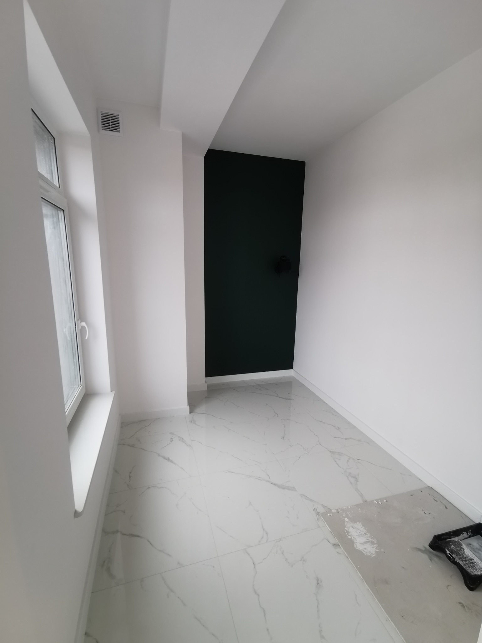 Wnętrze po odświeżeniu: białe ściany, ciemnozielona ściana akcentująca, marmurowe płytki podłogowe, okno z białymi ramami, widoczne ślady po malowaniu na podłodze i kuweta malarska.