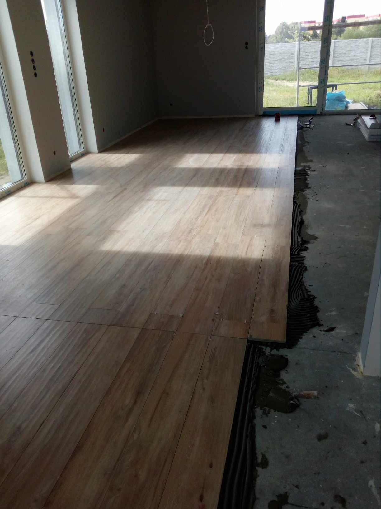 Układanie drewnopodobnych płytek podłogowych w jasnym pomieszczeniu z dużymi oknami, widoczny proces klejenia i czarny podkład izolacyjny.