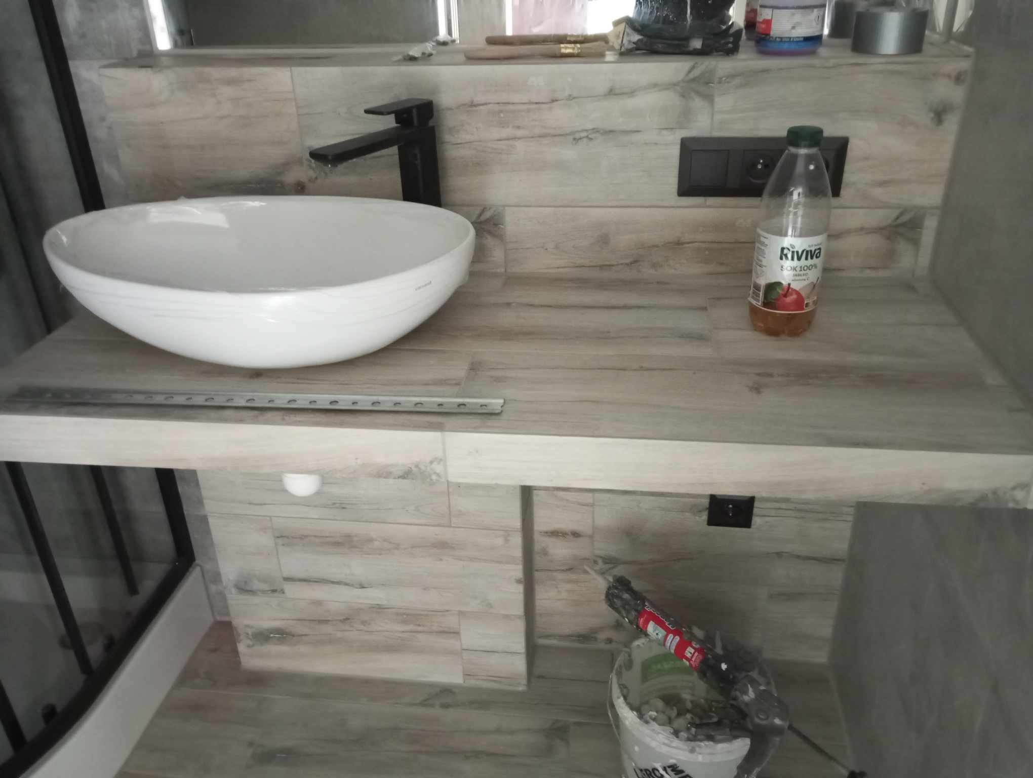 Nowoczesna łazienka w trakcie wykańczania: biała, okrągła umywalka nablatowa z czarną baterią na blacie z płytek imitujących drewno, widoczne narzędzia i materiały budowlane, butelka soku.