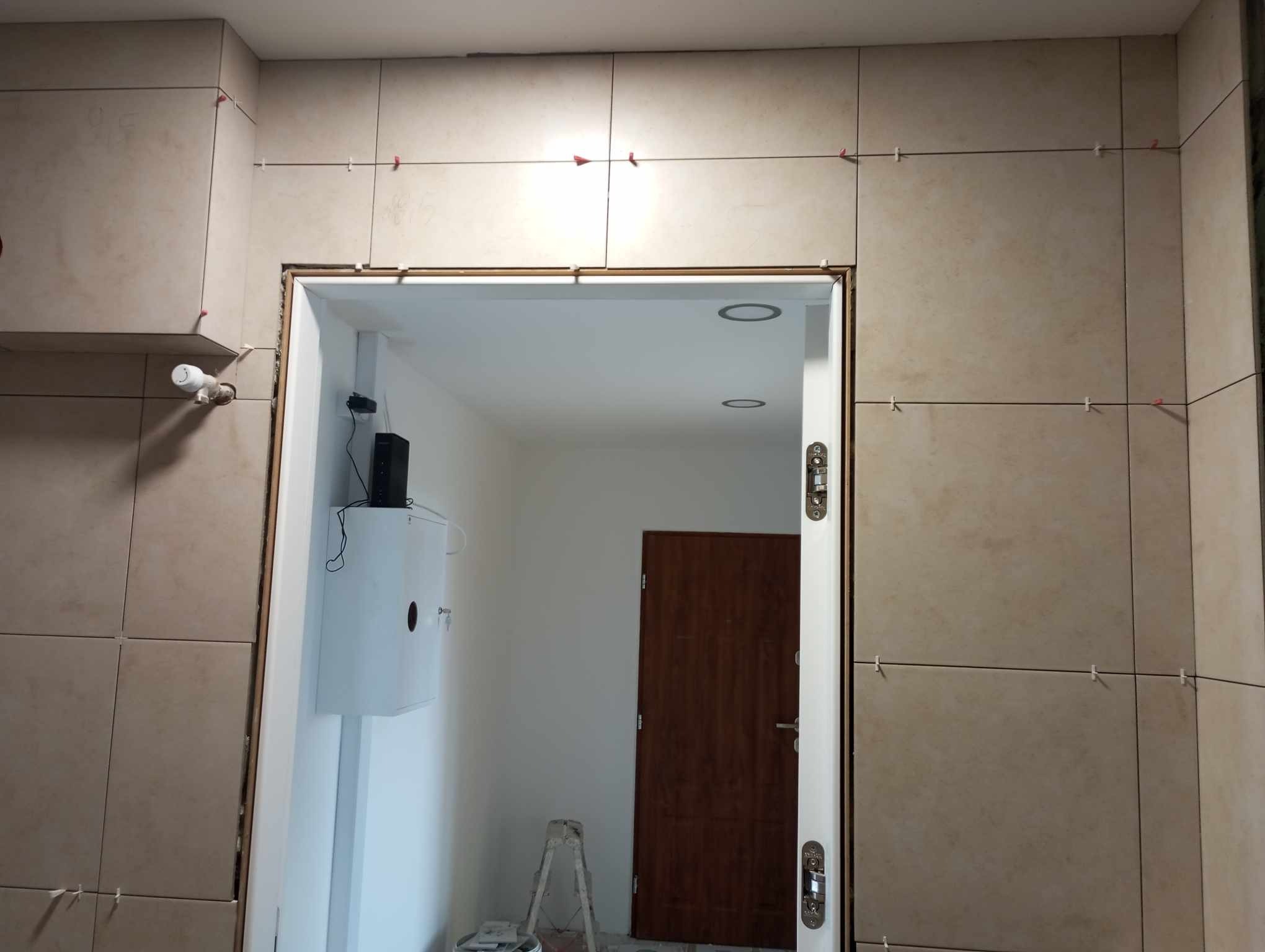 Układanie beżowych płytek w łazience, widoczne krzyżyki dystansowe, biała skrzynka elektryczna na ścianie i fragment drabiny malarskiej w przejściu do drugiego pomieszczenia.