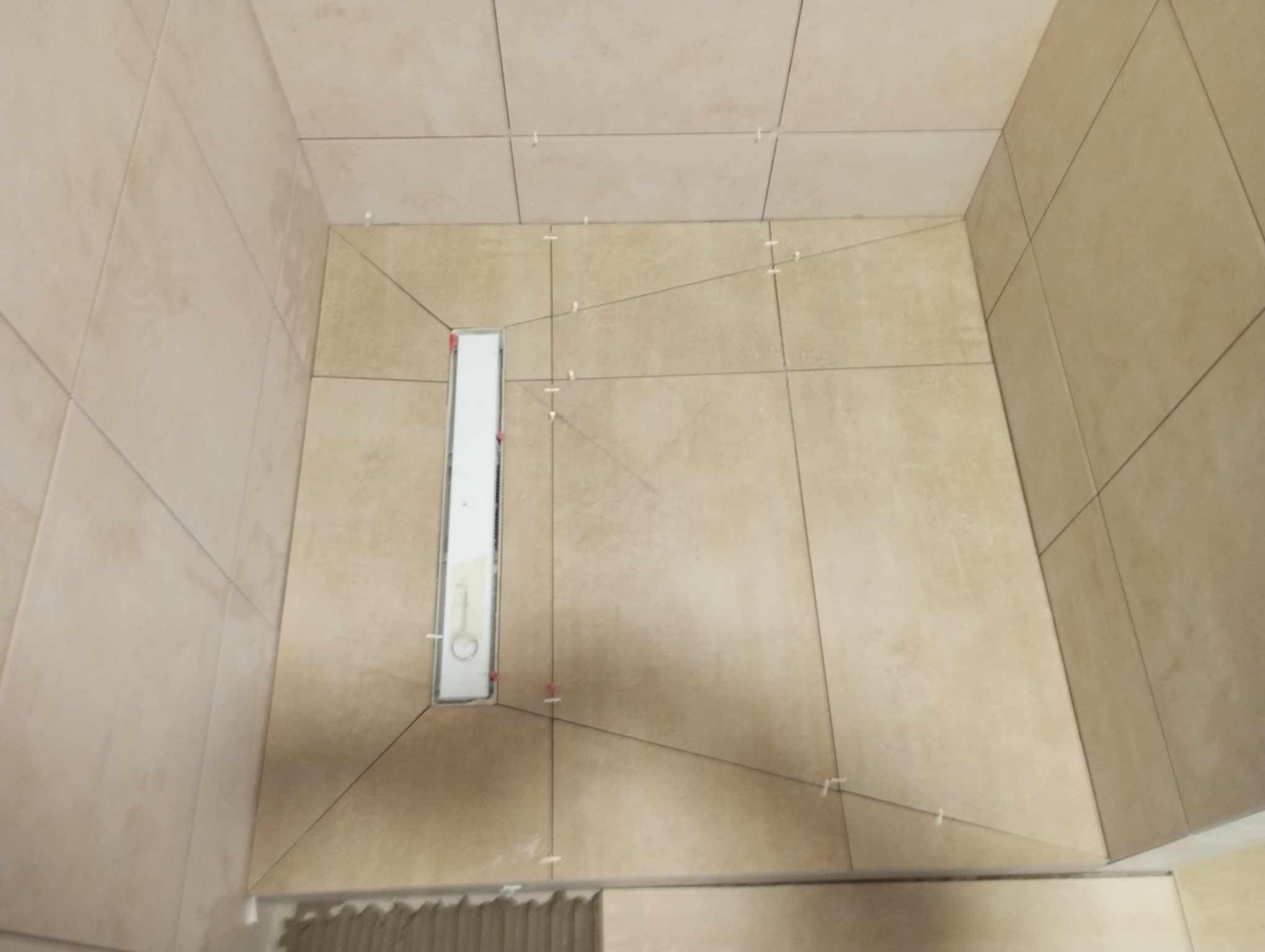 Beżowe płytki podłogowe i ścienne w trakcie układania w kabinie prysznicowej, widoczny odpływ liniowy i krzyżyki dystansowe.