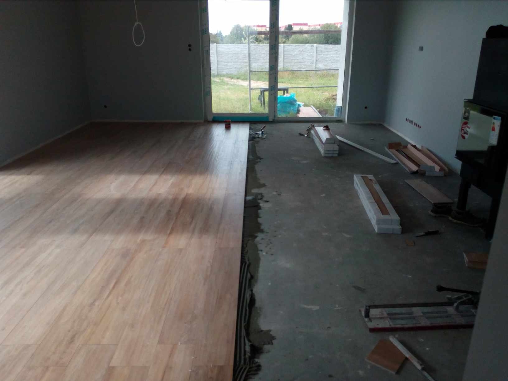 Układanie jasnych paneli podłogowych w surowym wnętrzu z widocznym klejem i narzędziami, częściowo wykończona podłoga, okno balkonowe z widokiem na podwórze.