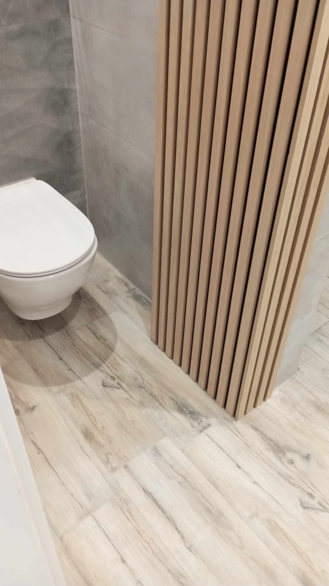 Wykończona łazienka z toaletą, płytkami imitującymi drewno na podłodze i ścianie oraz dekoracyjnymi panelami lamelowymi z jasnego drewna.