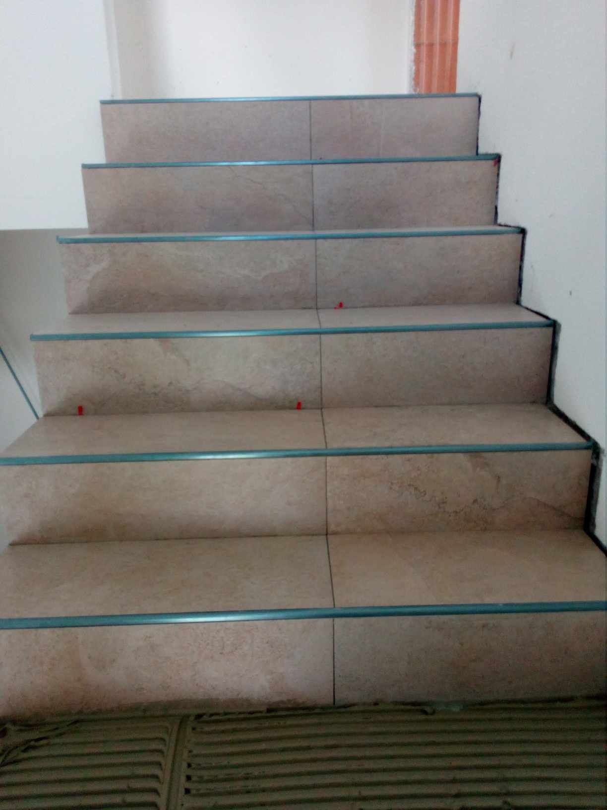 Wykończone schody wewnętrzne z beżowych płytek z niebieskimi listwami krawędziowymi, widoczne czerwone kliny poziomujące płytki.
