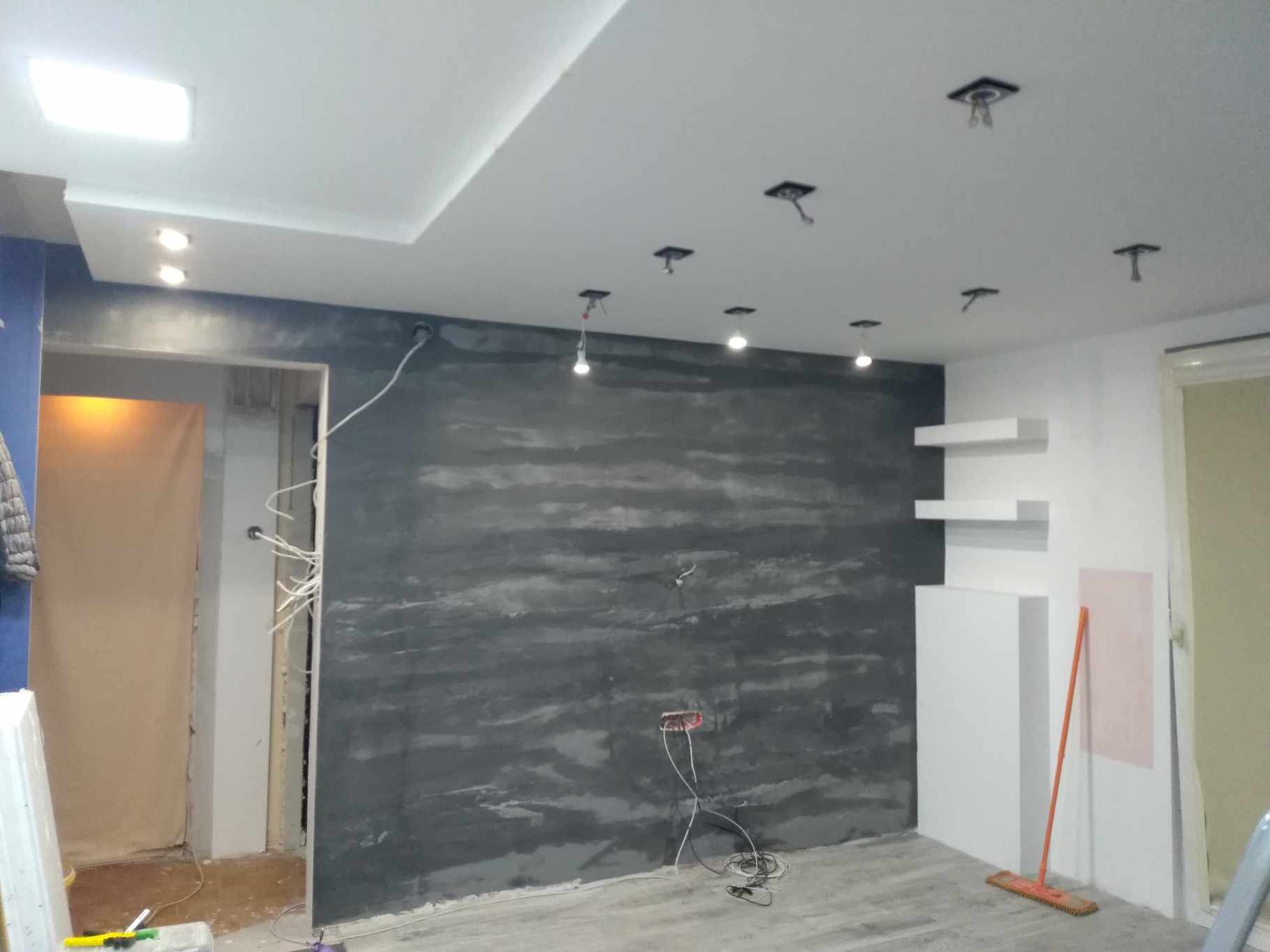 Wnętrze w trakcie prac wykończeniowych: ściana z efektem dekoracyjnym, sufit z instalacją oświetleniową, widoczne kable, półki ścienne i narzędzia sprzątające.