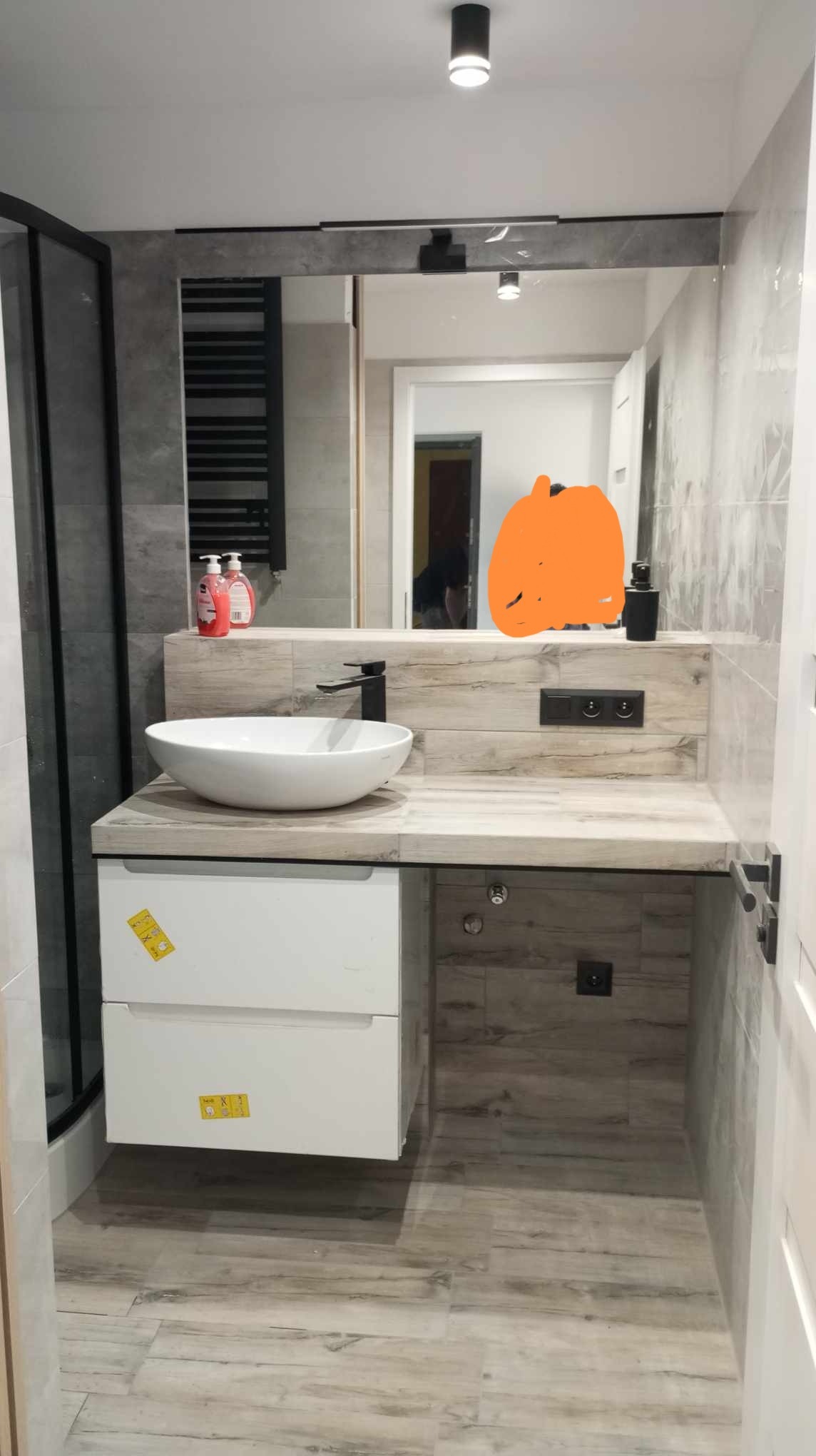 Nowoczesna łazienka z umywalką nablatową, czarną armaturą i blatem imitującym drewno, z widocznym grzejnikiem drabinkowym w tle i szklaną kabiną prysznicową po lewej stronie.