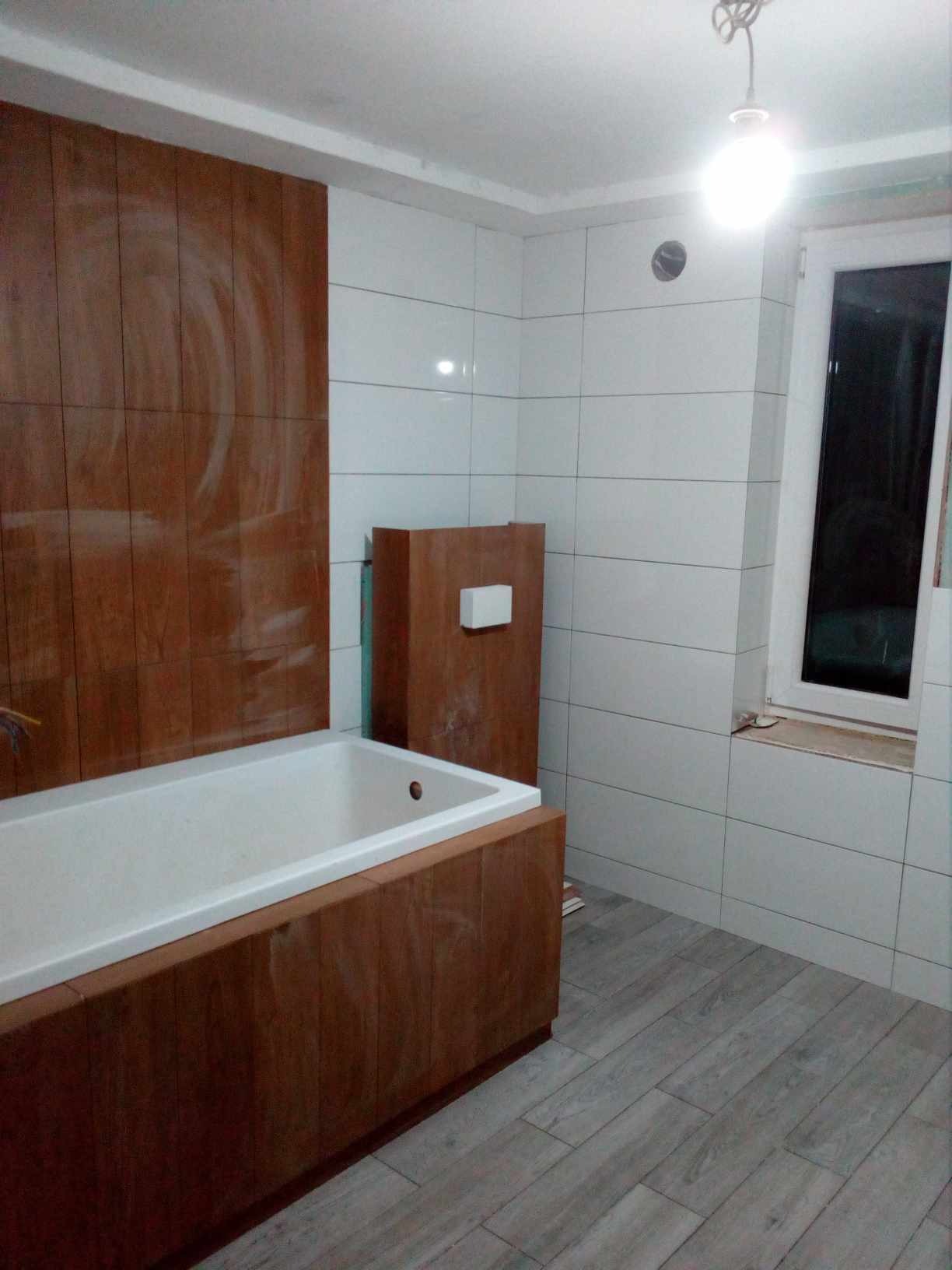 Wnętrze łazienki w trakcie wykańczania: białe płytki ścienne, drewnopodobna obudowa wanny i płyty ścienne, szare płytki podłogowe imitujące drewno, surowa żarówka zwisająca z sufitu, okno...